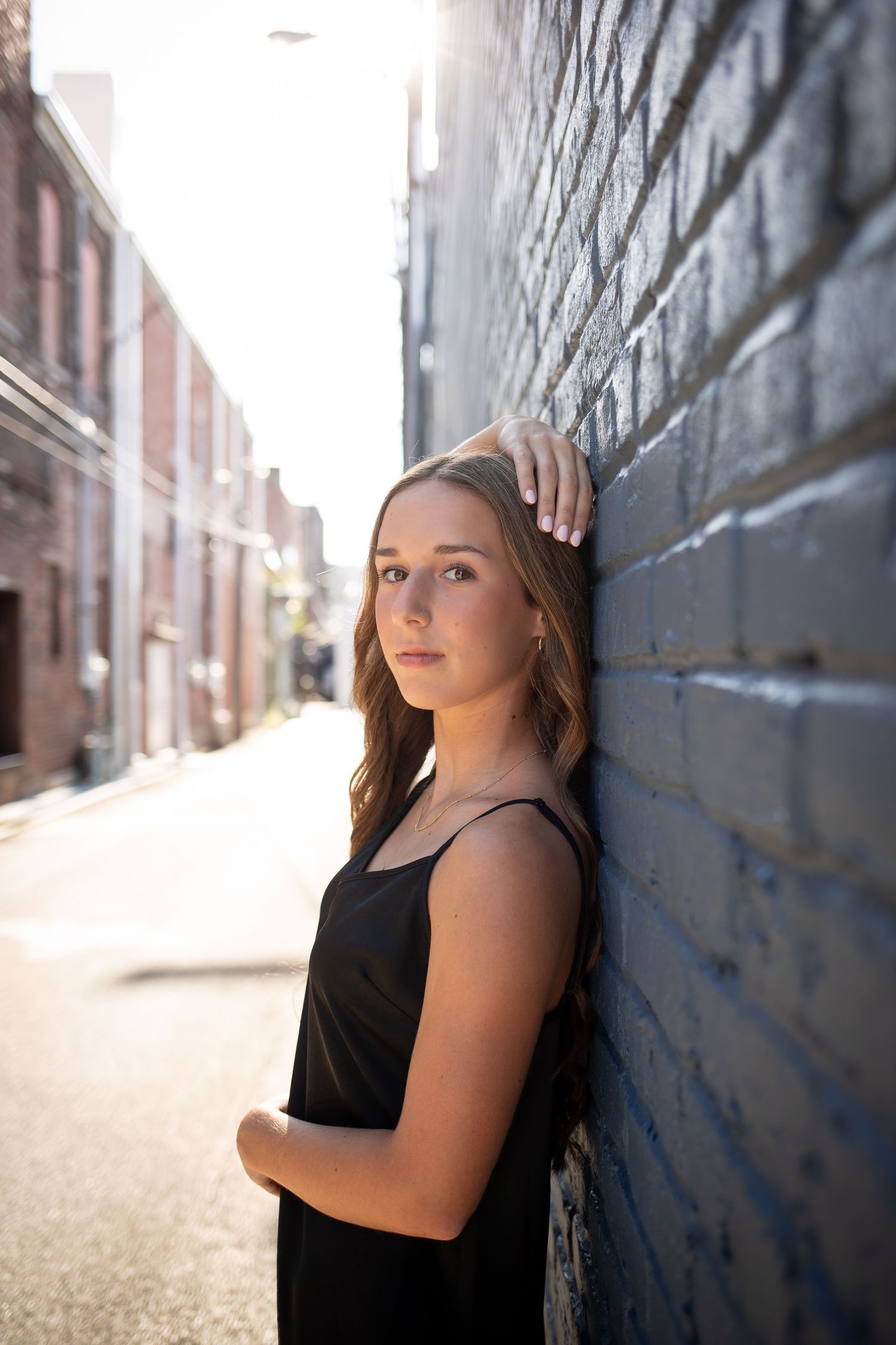 Portrait of someone in a black sleeveless top leaning against a dark brick wall in an urban alley with dramatic lighting.