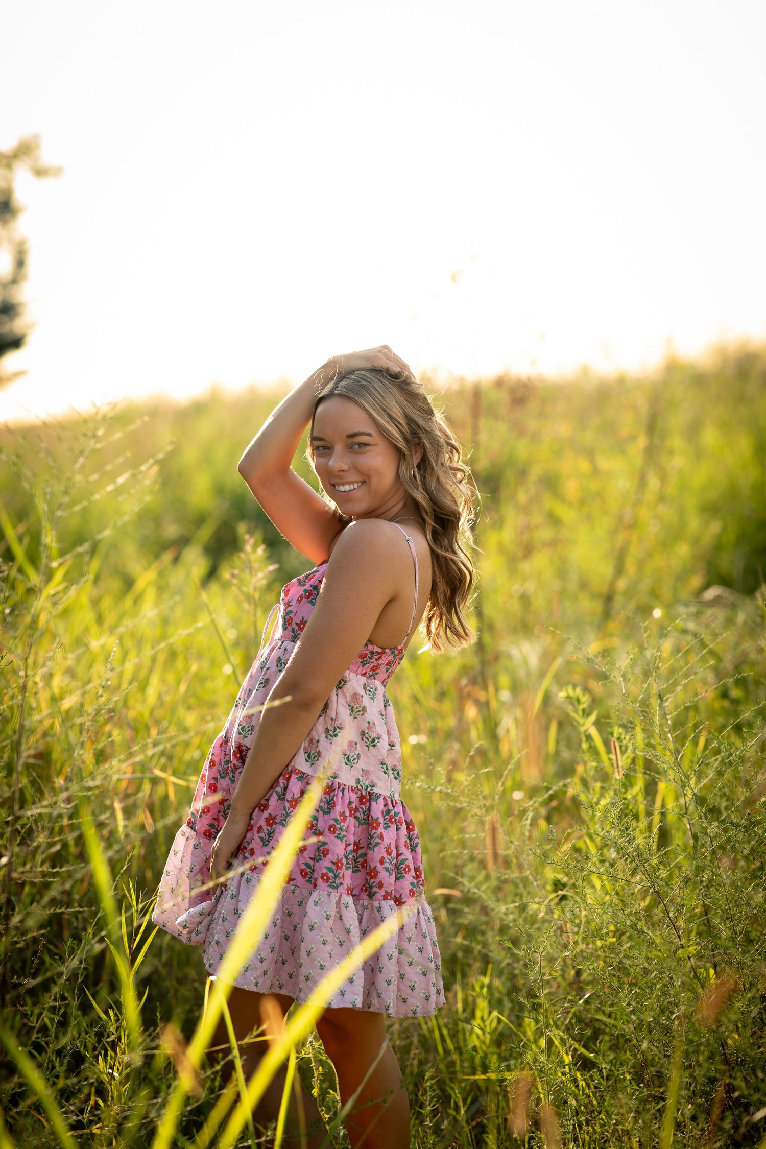 A person in a floral dress poses playfully in a sunlit meadow during golden hour.