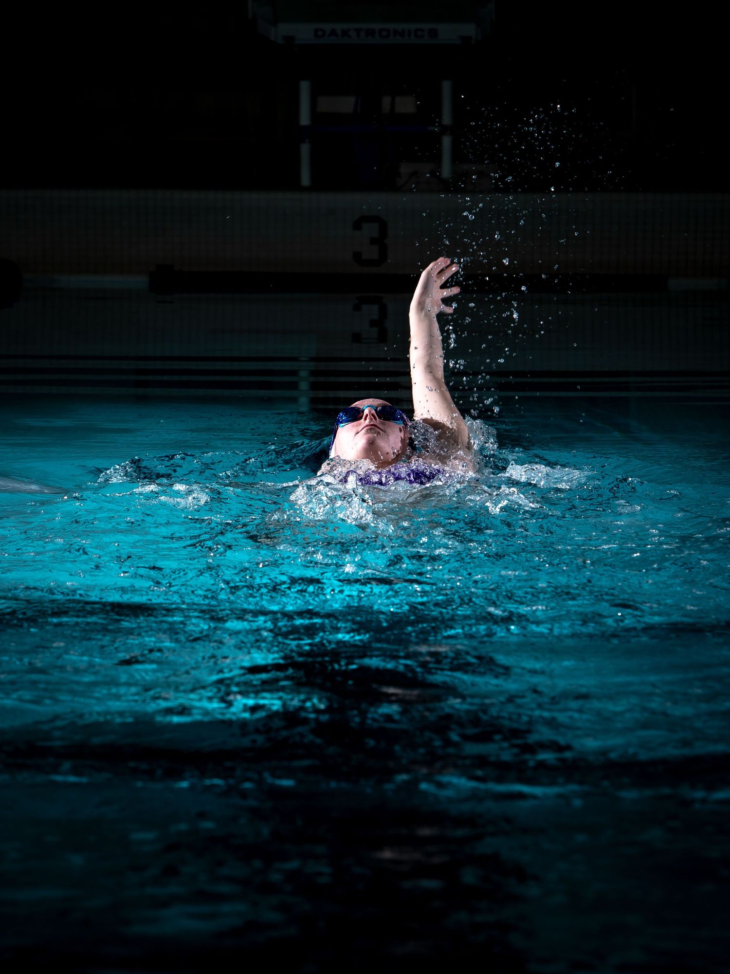 A swimmer performs a backstroke in a dimly lit indoor pool creating dynamic splashes in turquoise blue water.