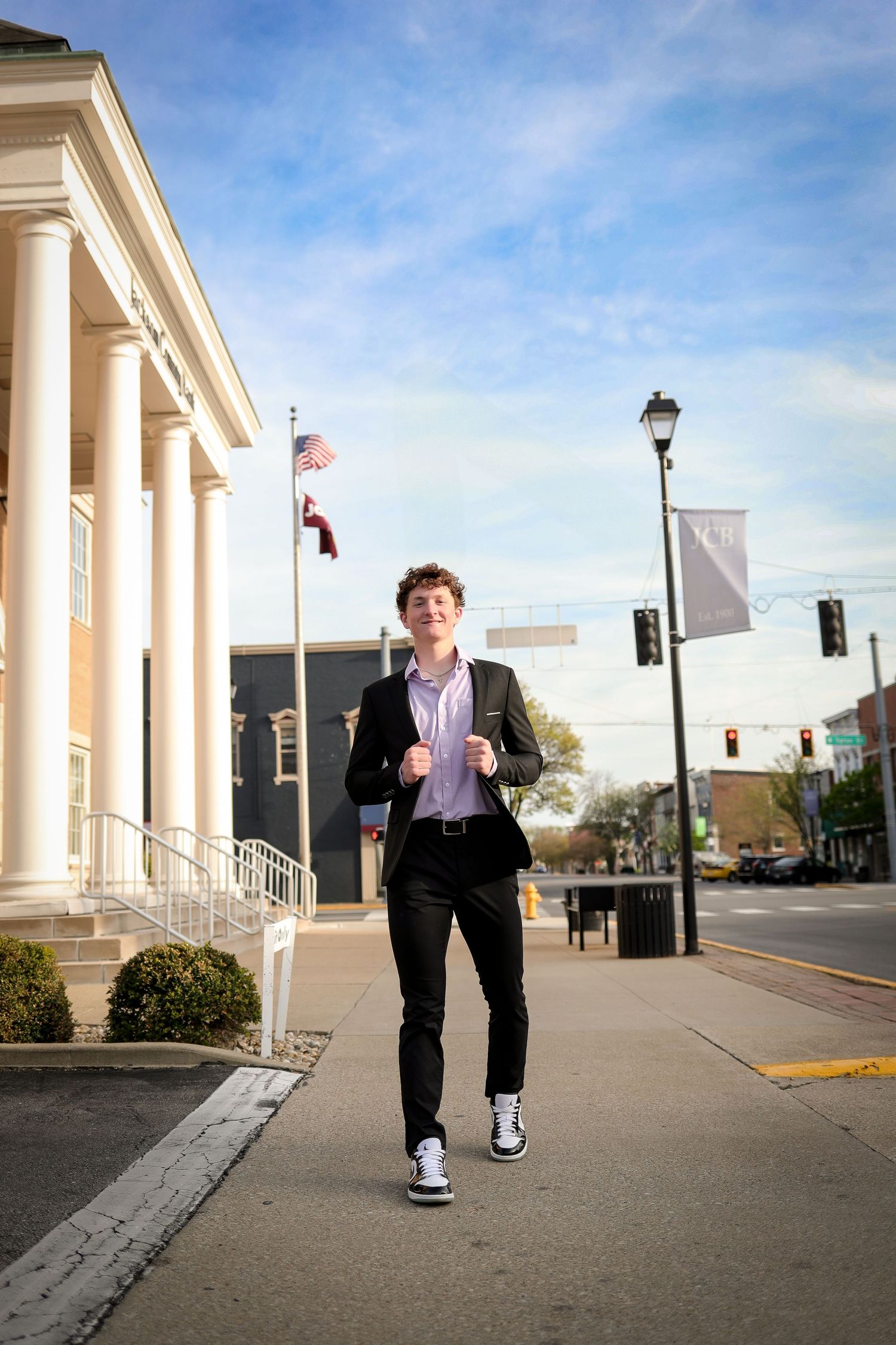 A person in a suit walks along a colonnade with white pillars against a blue sky and urban backdrop.