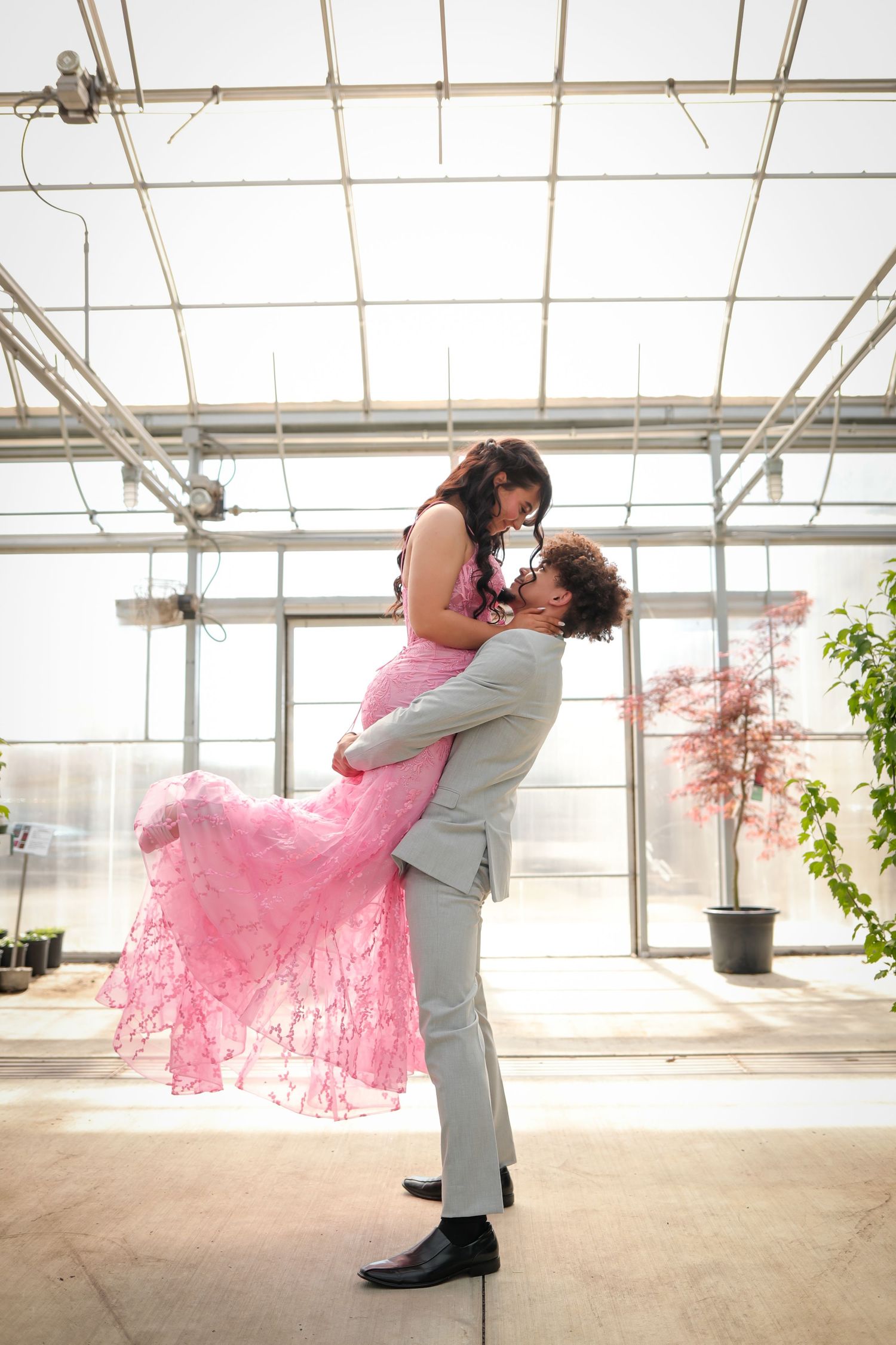 A couple in a greenhouse shares a romantic dance dip pose, with the girl wearing a flowing pink dress and boy in gray.