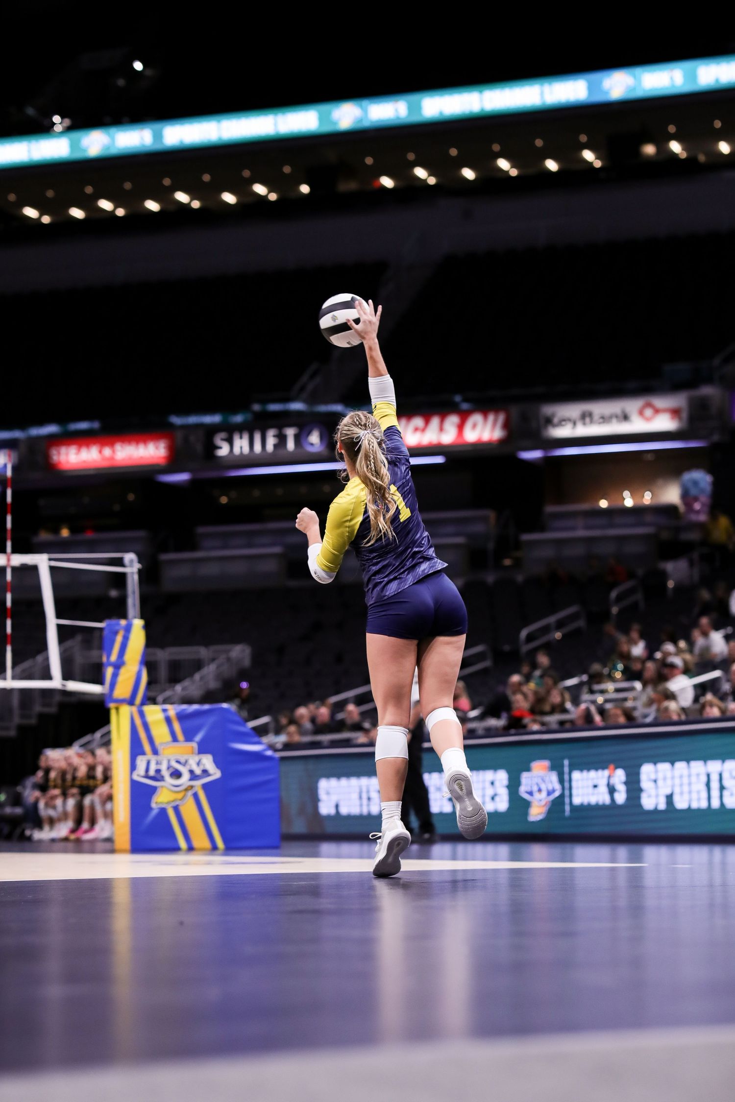 Female volleyball player in navy uniform jumps high to serve the ball during indoor match on professional court.
