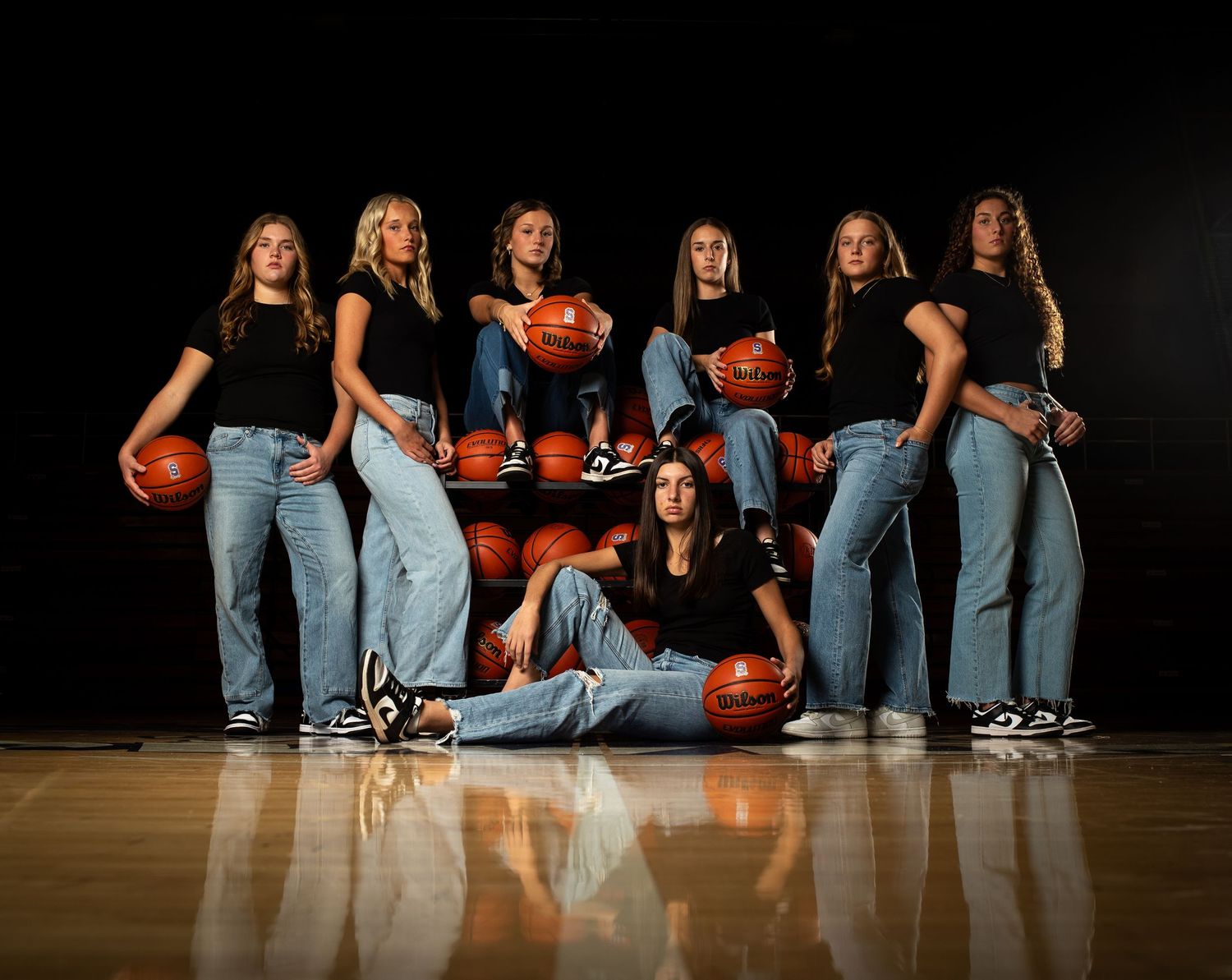 Basketball players in blue jeans sit on a wooden court floor holding basketballs in a creative team photo composition.