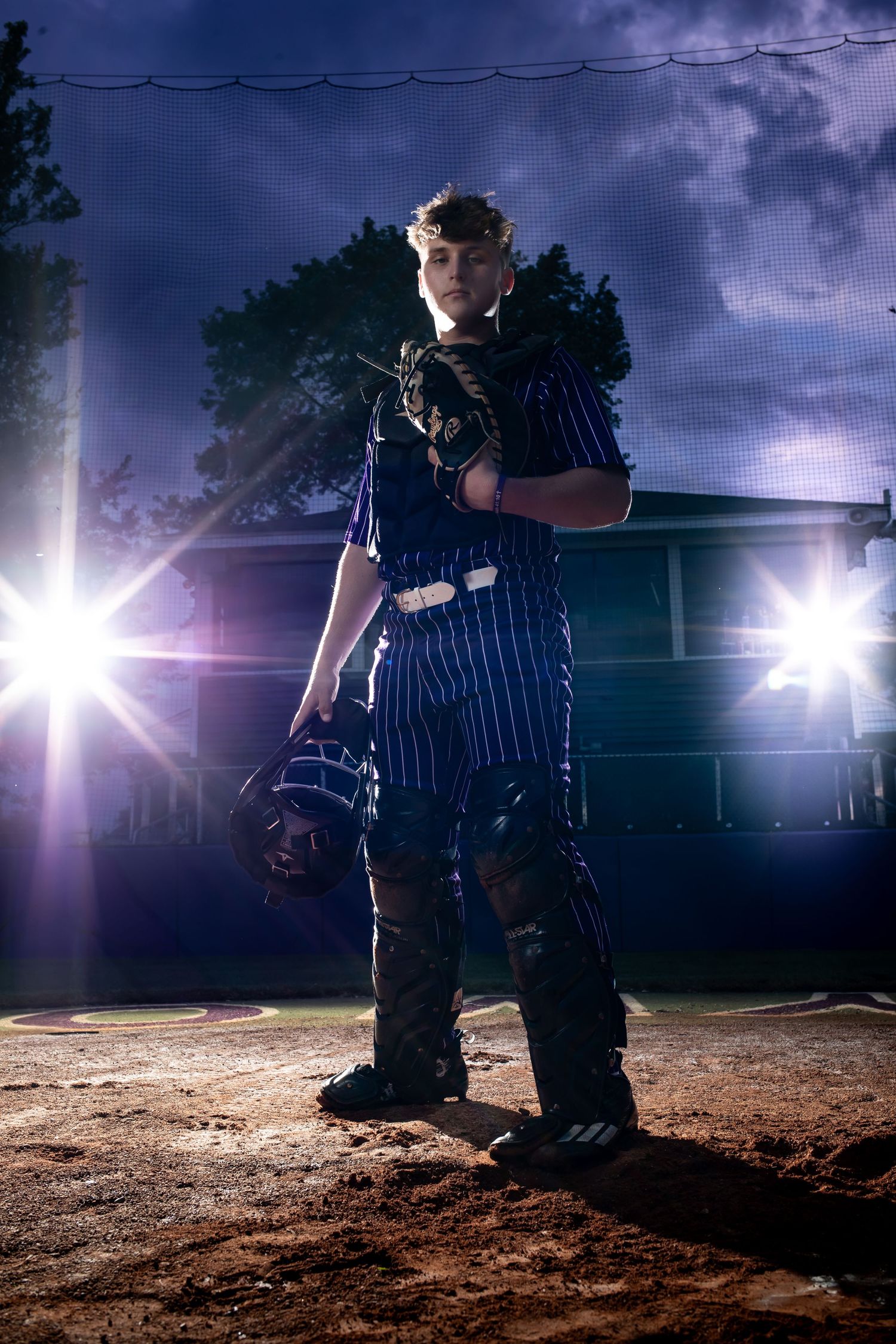 Baseball player in pinstripe uniform stands on dirt field at night with dramatic stadium lighting creating silhouette effect.