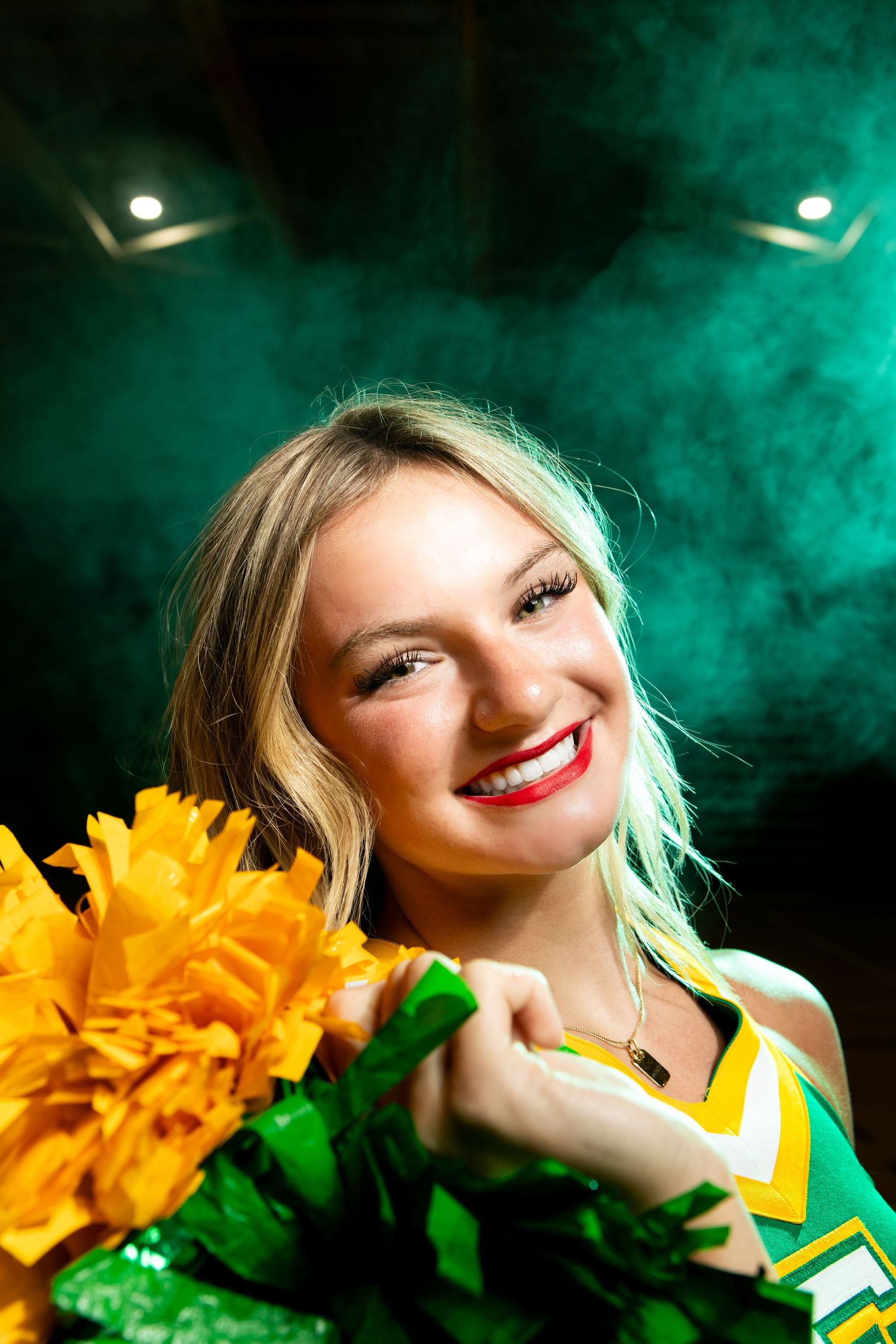 Cheerleader in green and yellow uniform holds bright yellow pom poms against emerald smoke background.