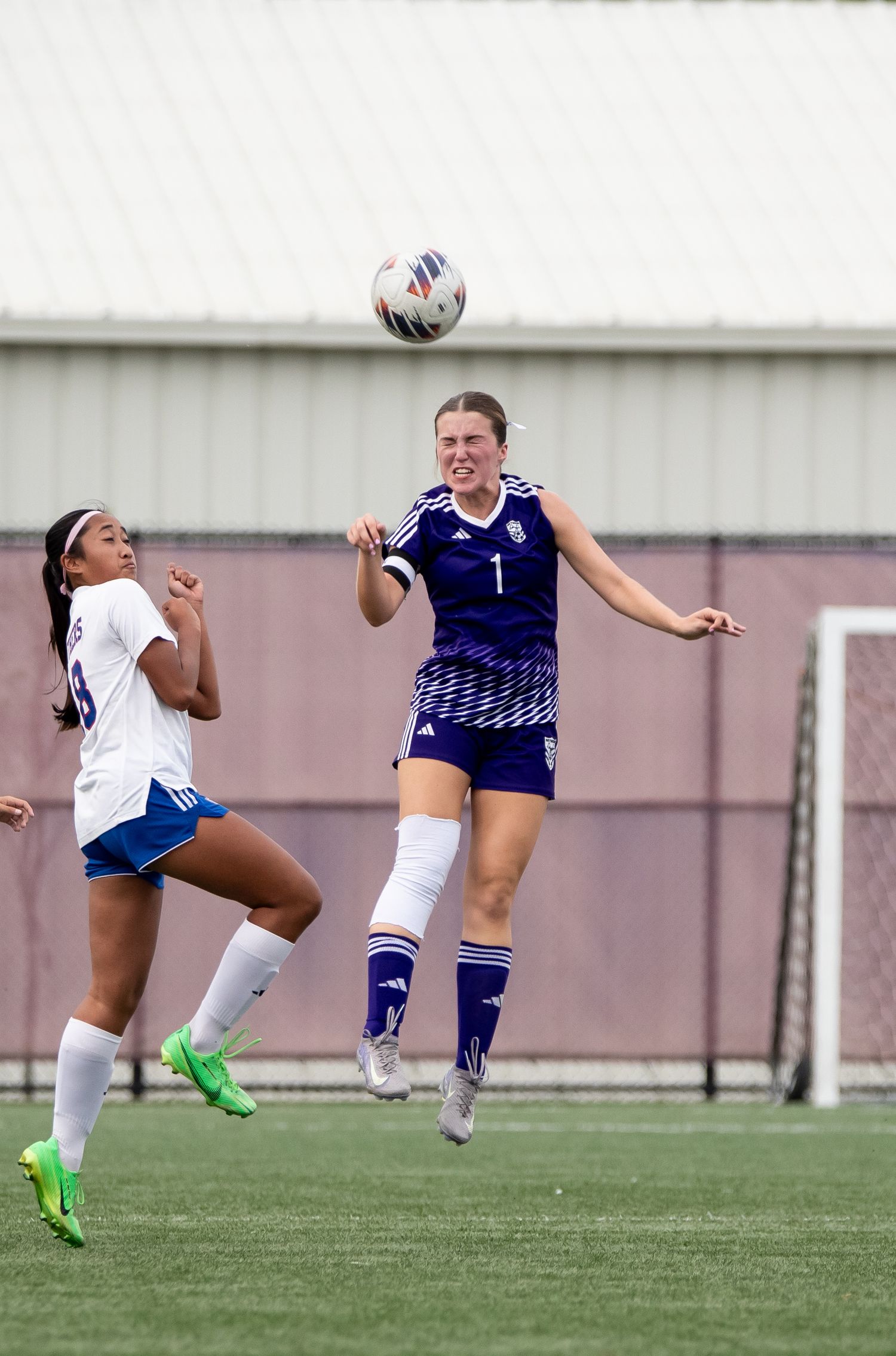 Two soccer players leap into the air to compete for a header during an intense match on an artificial turf field.