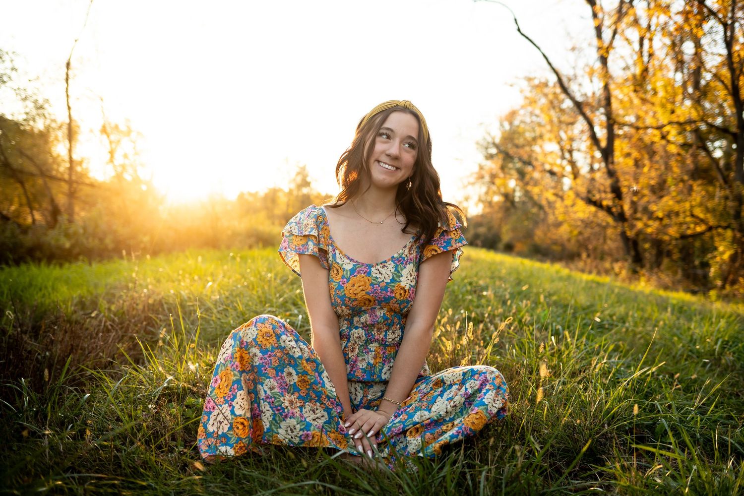 A woman in a floral dress sits in a sunlit meadow during golden hour with autumn trees in the background.