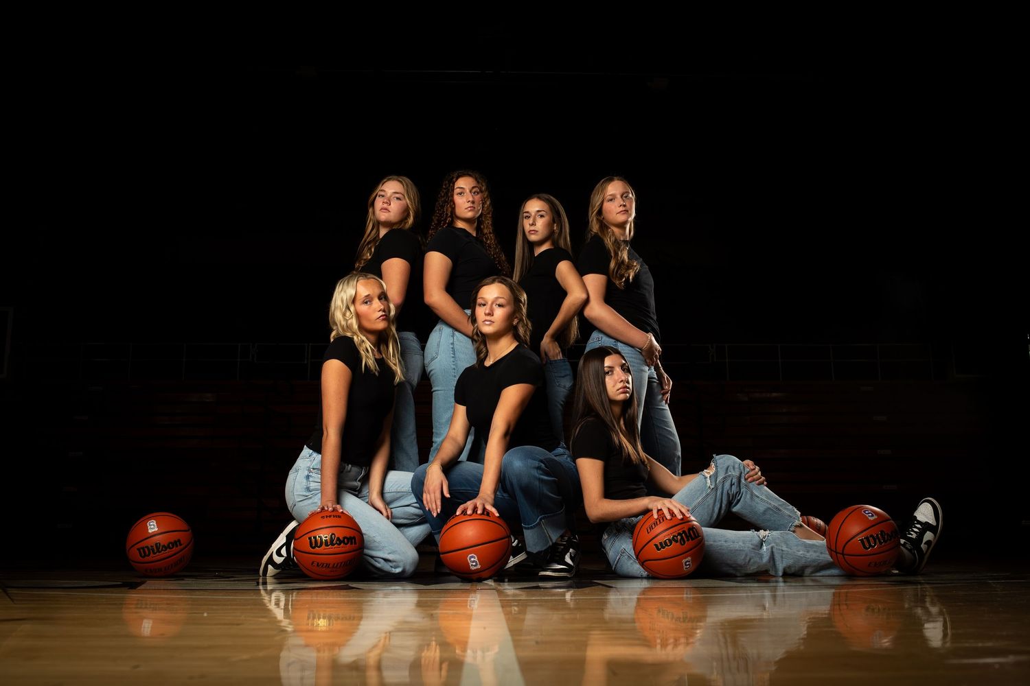 A group of basketball players in matching uniforms pose with orange basketballs on a dark wooden court.