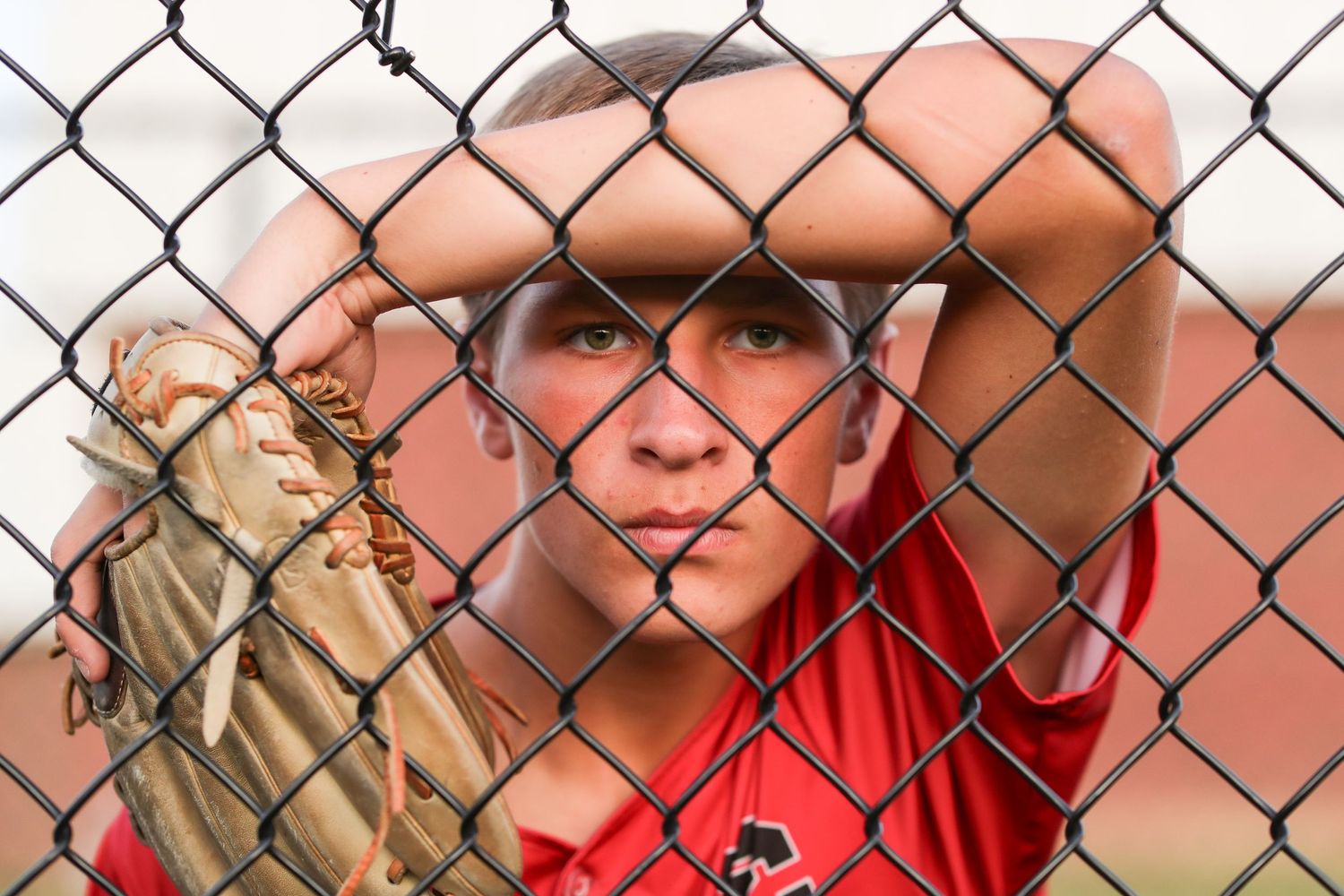 A baseball player in a red uniform watches through a chain link fence during a game.