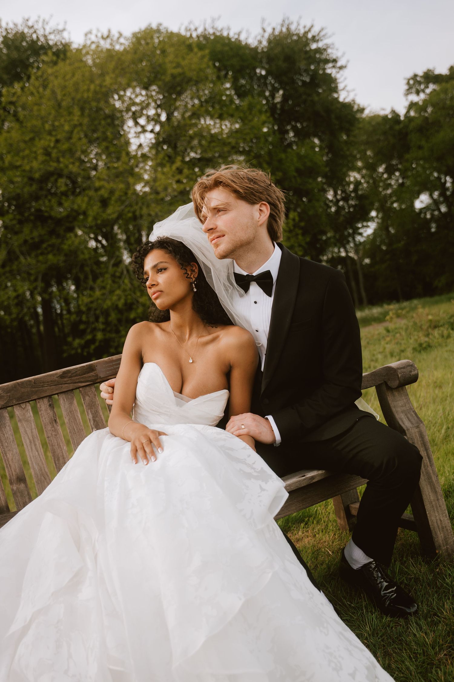 A bride and groom sit together on a wooden bench outdoors, surrounded by lush green trees on their wedding day.