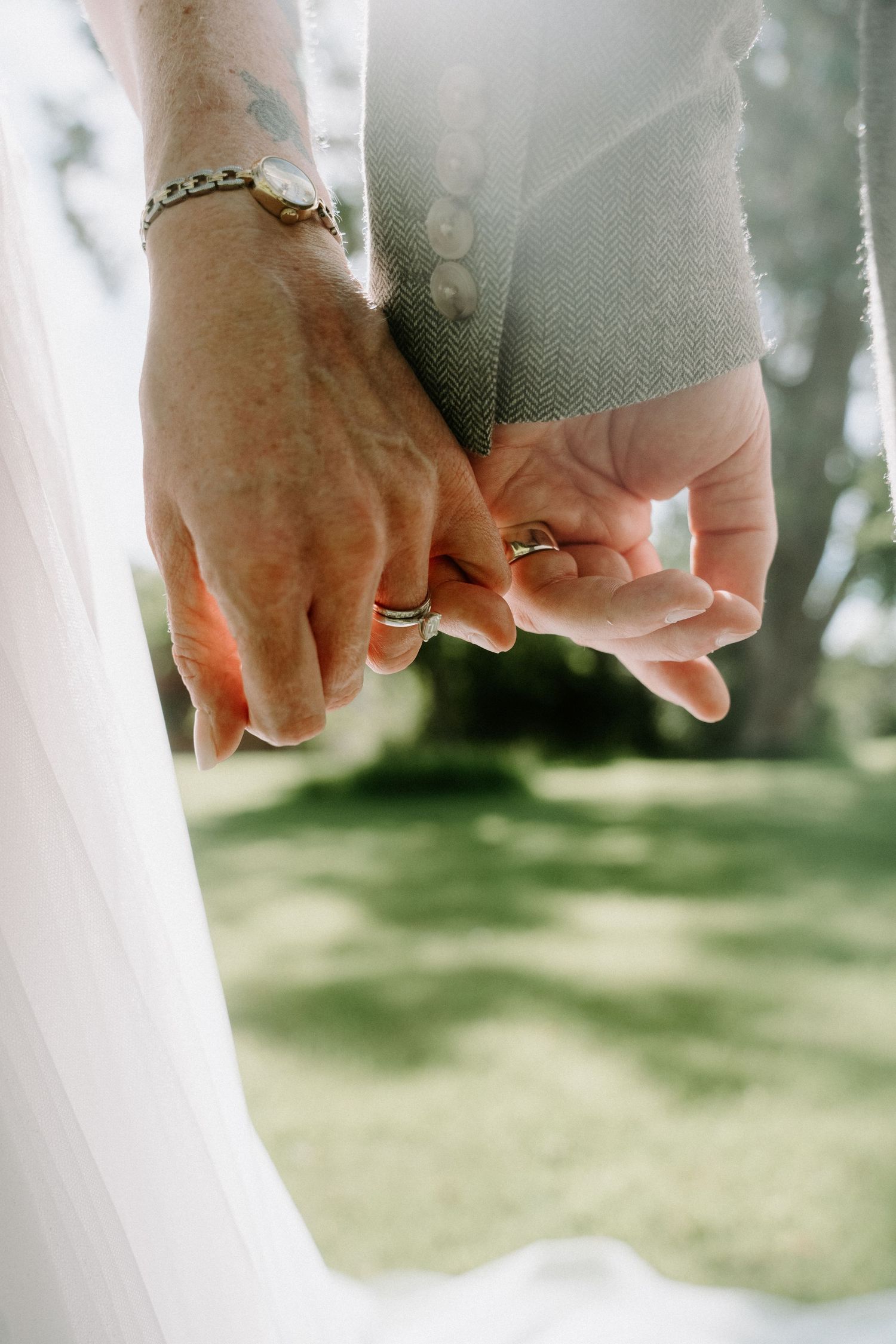Elderly couple holding hands outdoors, wearing wedding rings, with a white veil and green trees in the background.