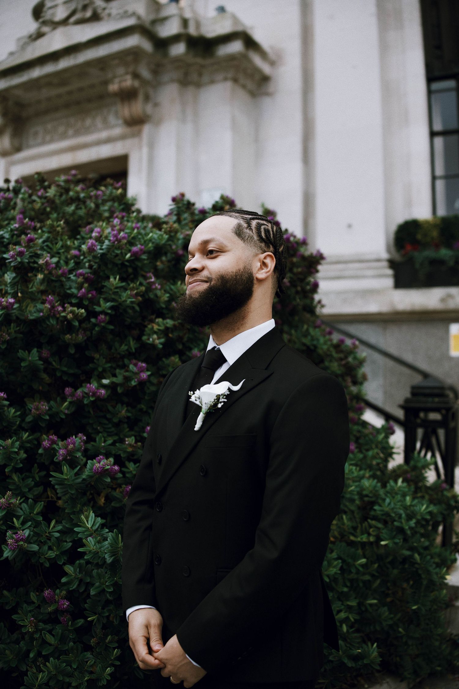 A well-dressed groom in a black suit with boutonniere smiles outdoors near lush green bushes and a classic building.