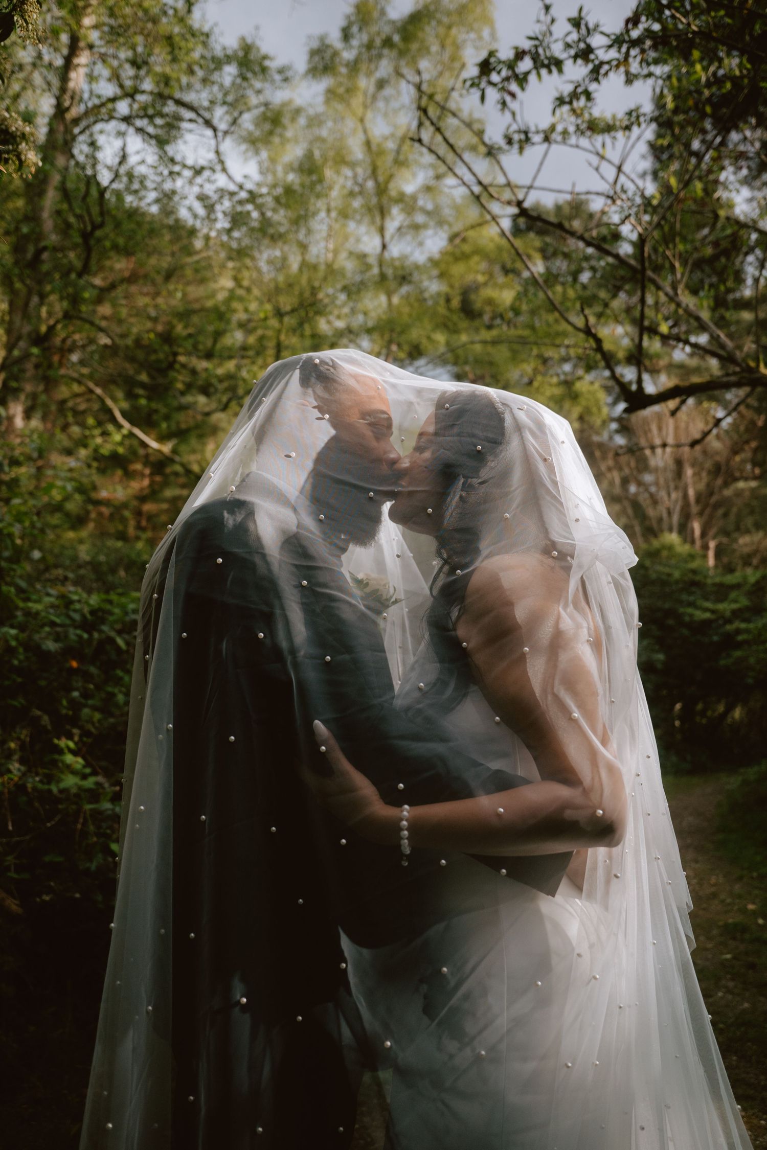 Bride and groom sharing a romantic kiss under a pearl-dotted wedding veil in a lush green garden setting.