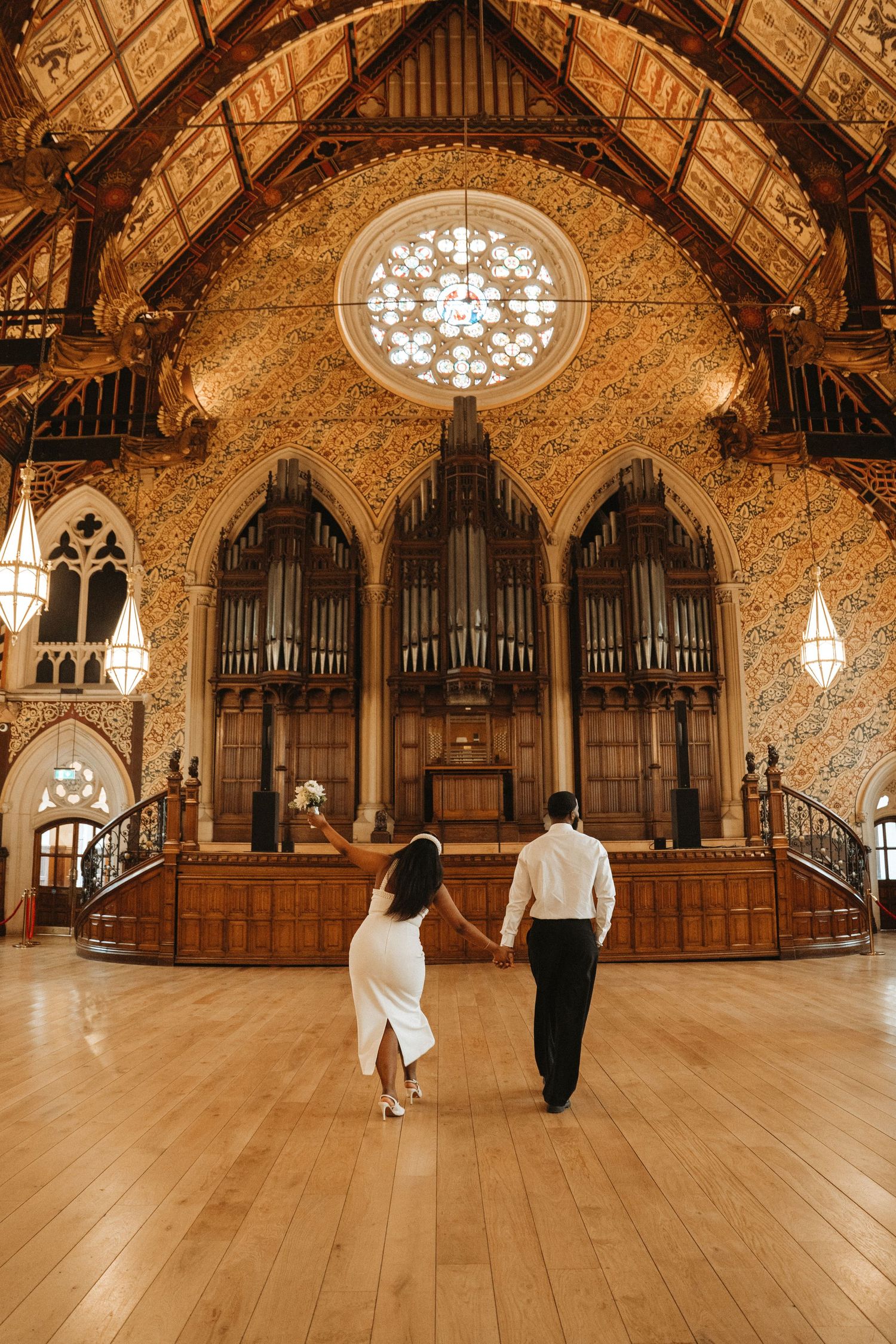 Couple holding hands walking toward ornate church organ under rose window on polished wood floor.