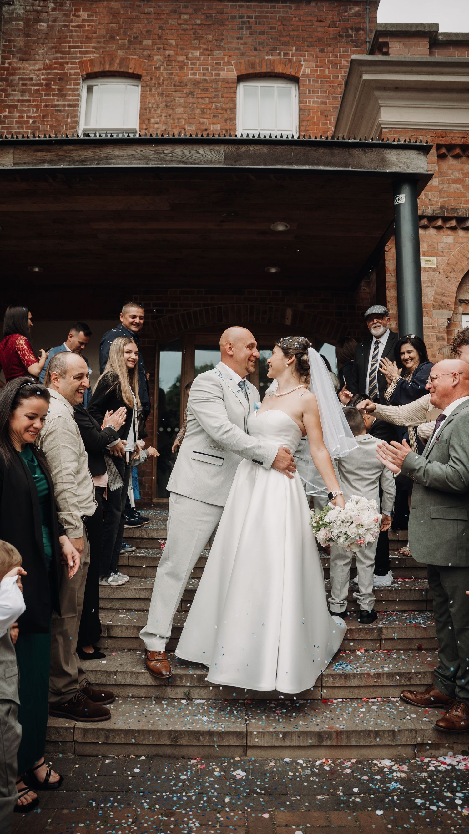Wedding celebration with confetti outside a historic brick building as guests cheer.
