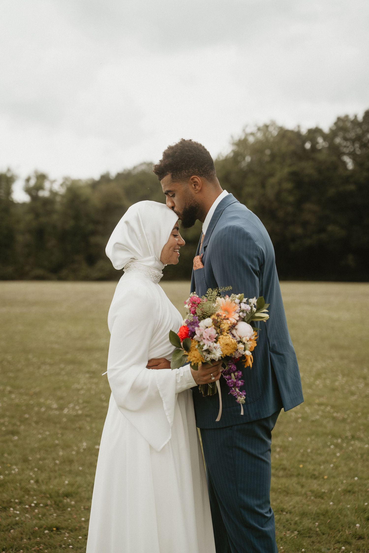 Wedding couple in field holding colorful bouquet against cloudy sky.