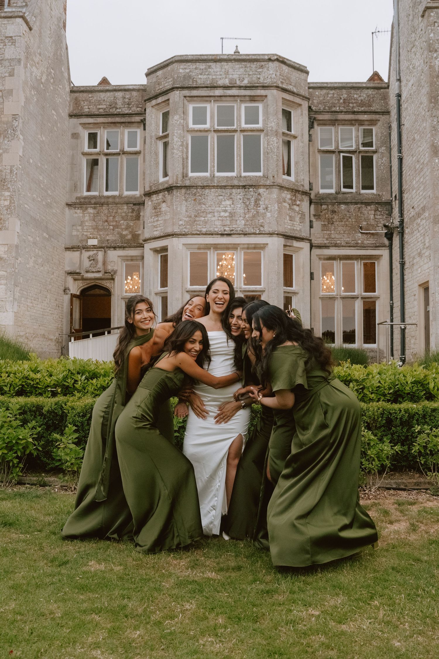 Bride in white gown surrounded by bridesmaids in olive green dresses, laughing together outside a grand stone manor.