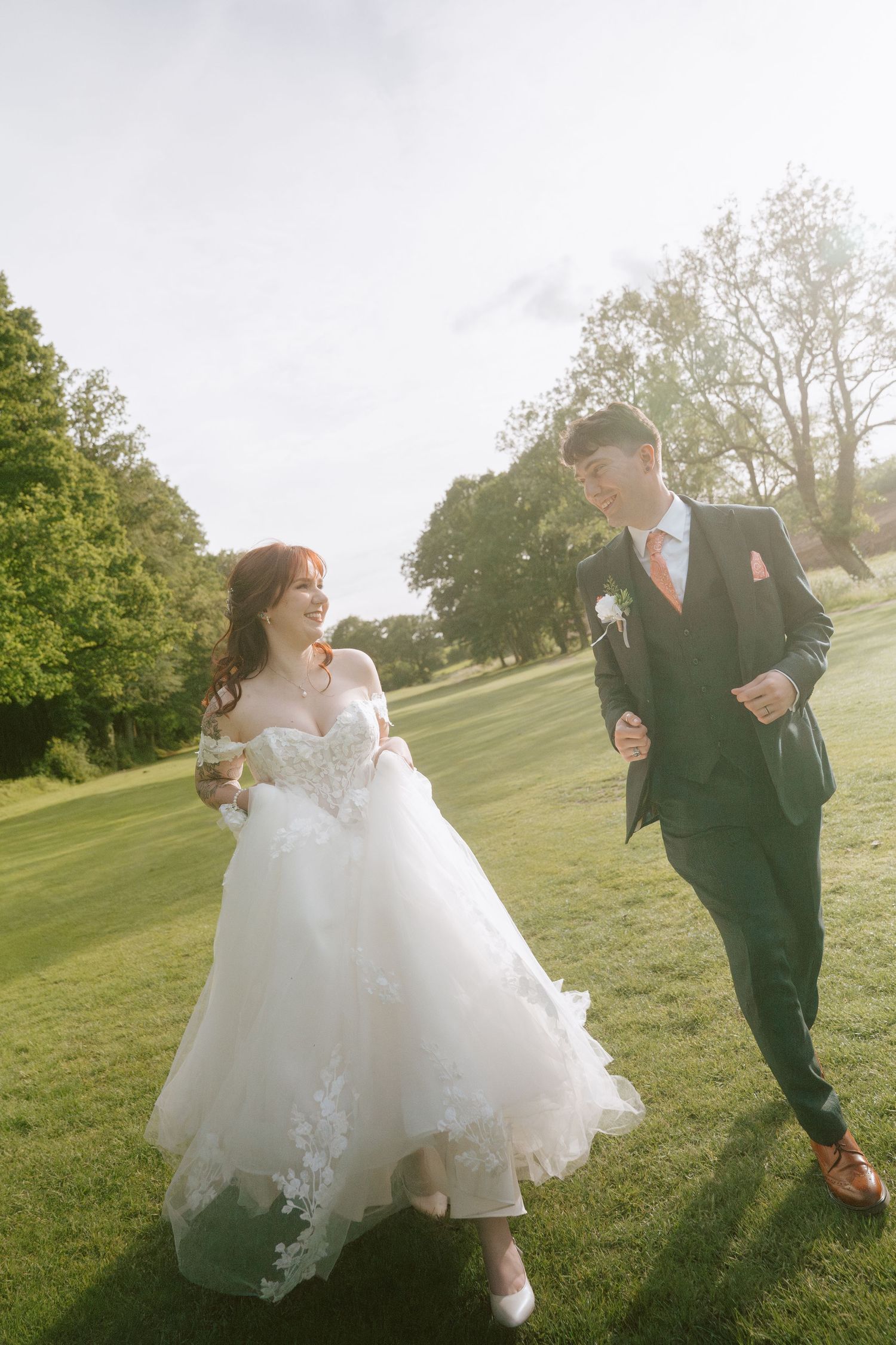 Bride and groom walking hand in hand on lush green lawn in golden sunlight, bride in lace ballgown, groom in navy suit.