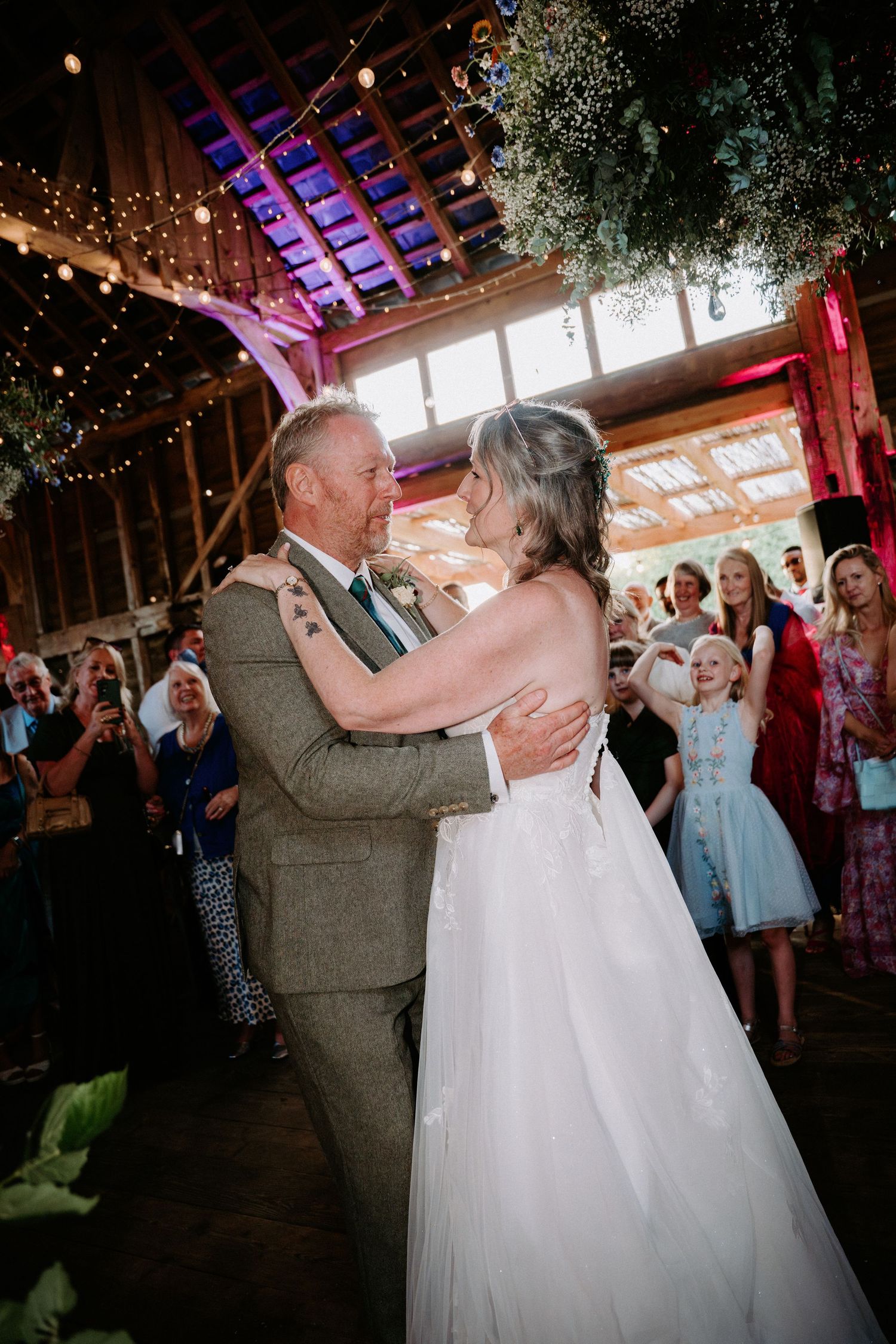 Bride and groom share their first dance in a rustic barn wedding venue with fairy lights and guests watching.