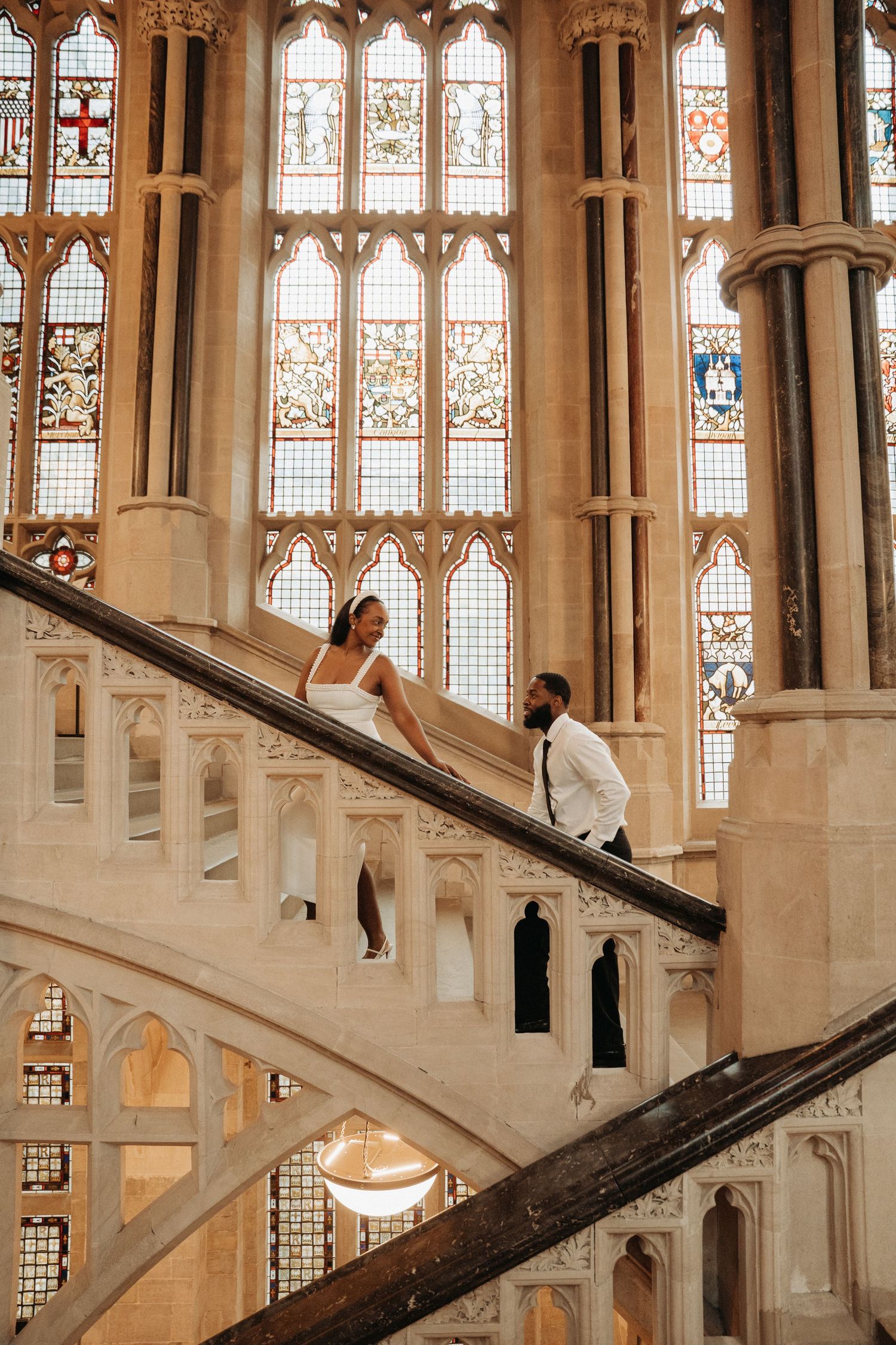 People walk up and down an ornate Gothic staircase with stained glass windows in a historic building interior.