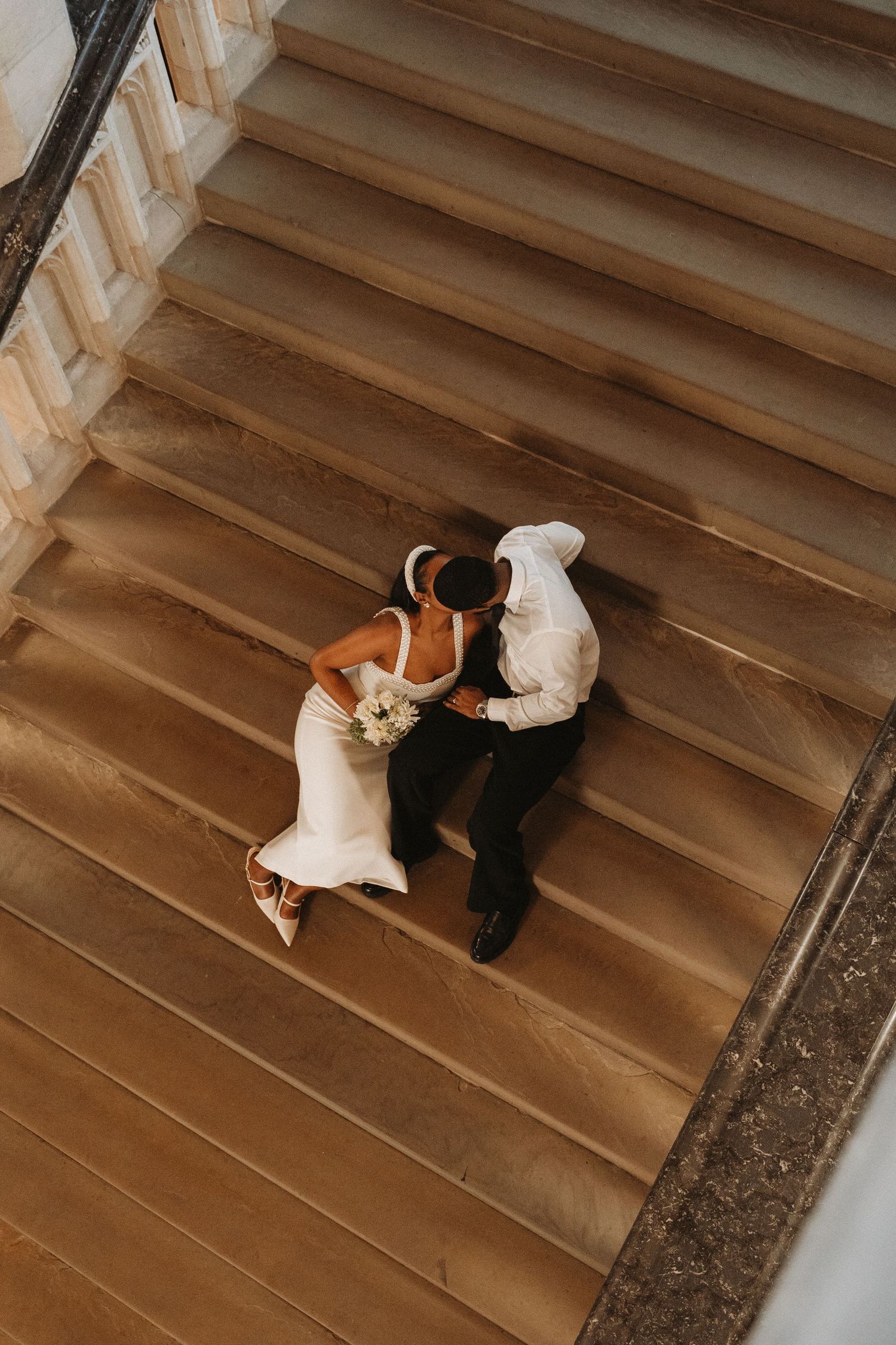 Aerial view of bride and groom kissing on grand staircase, bride holding bouquet in elegant white gown.