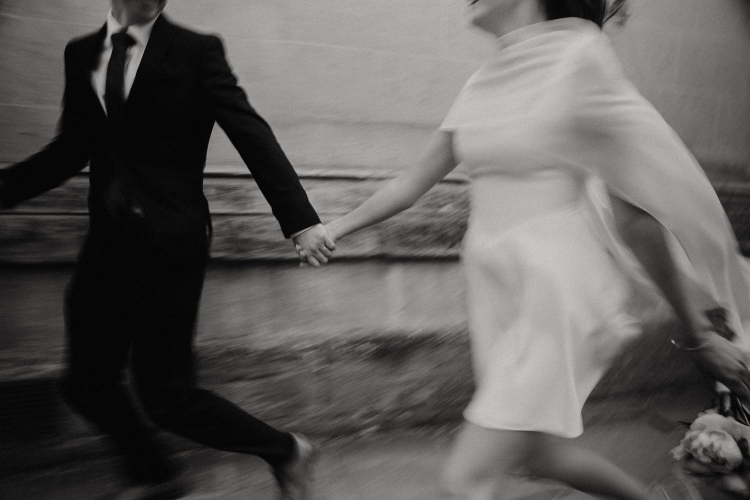 Black and white photo of a bride and groom holding hands and running together on their wedding day.