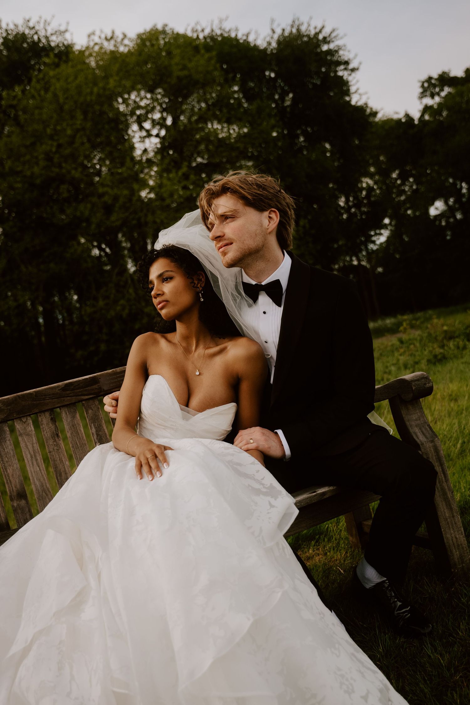Bride in white strapless gown and groom in black tuxedo sit on wooden bench surrounded by lush green trees.