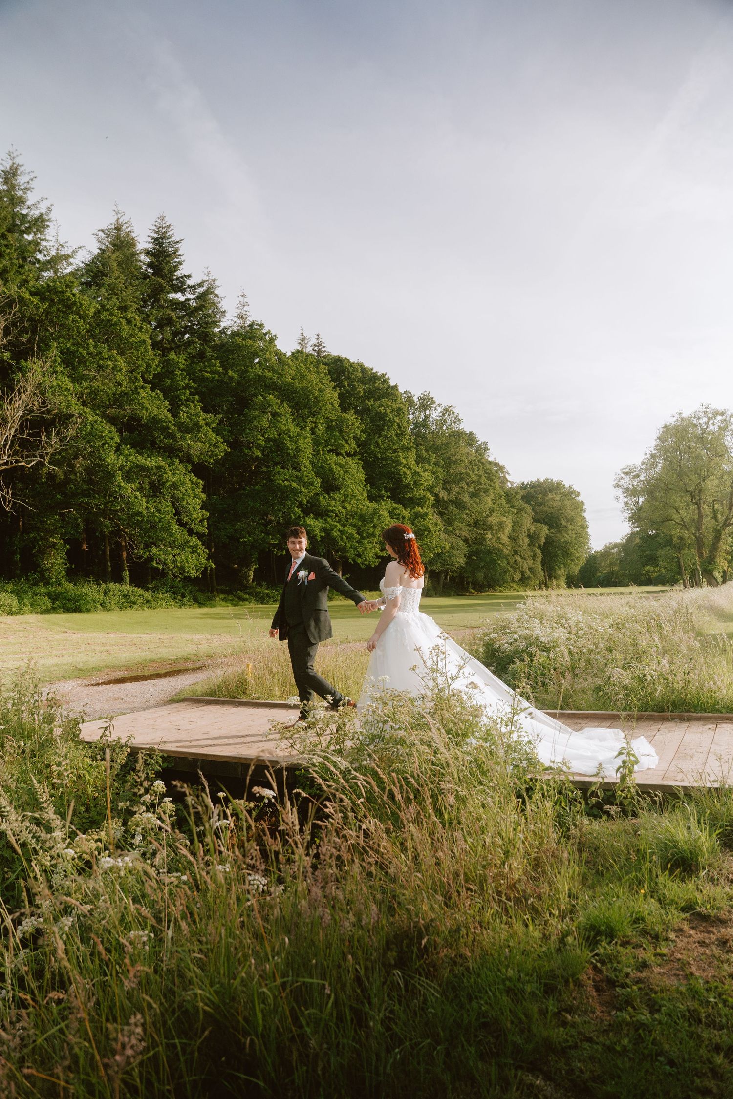 Bride and groom holding hands walking on wooden path surrounded by lush green trees and wildflowers on their wedding day.