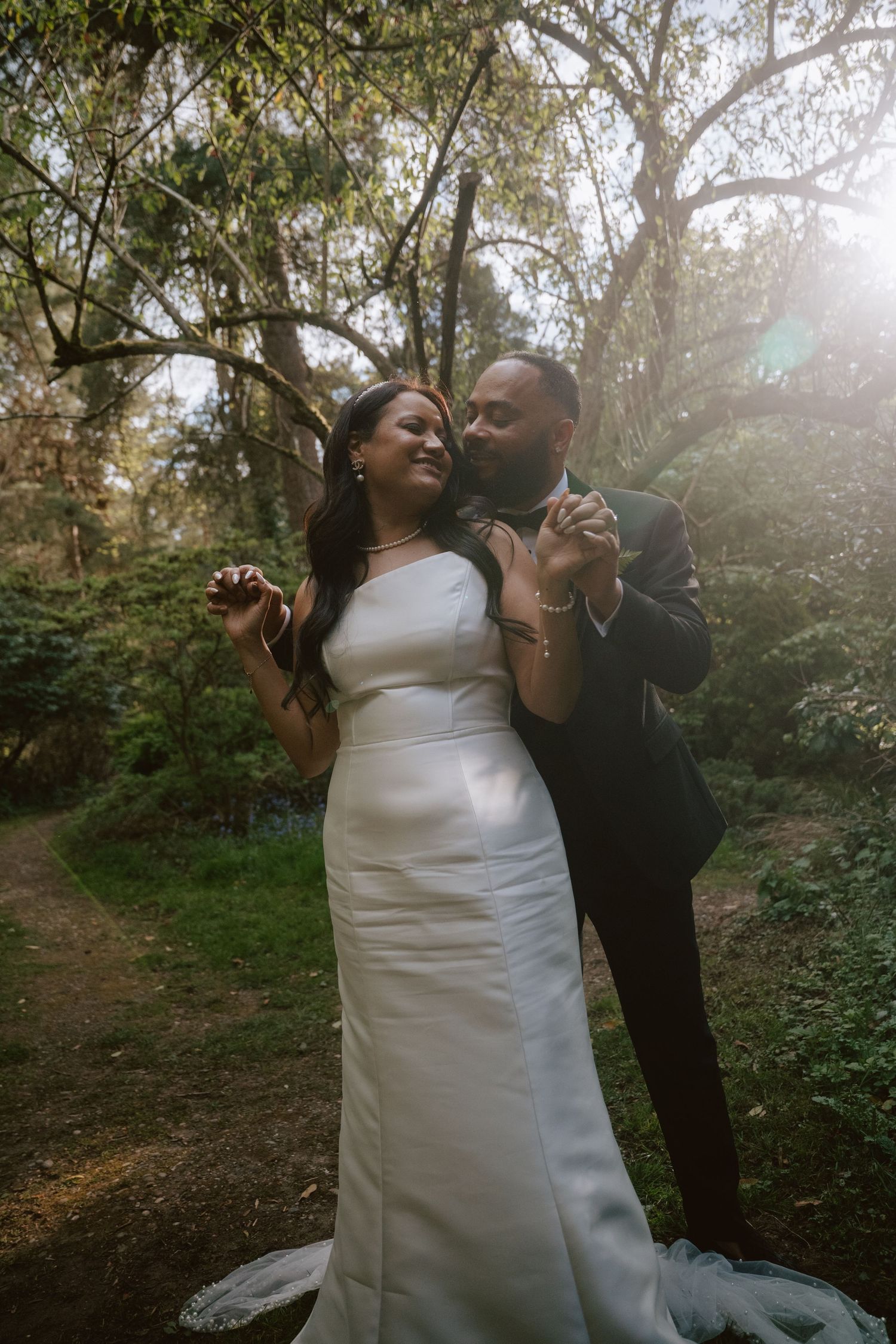 A bride and groom share a romantic dance in a sunlit forest, the bride in a sleek white gown, groom in a dark suit.