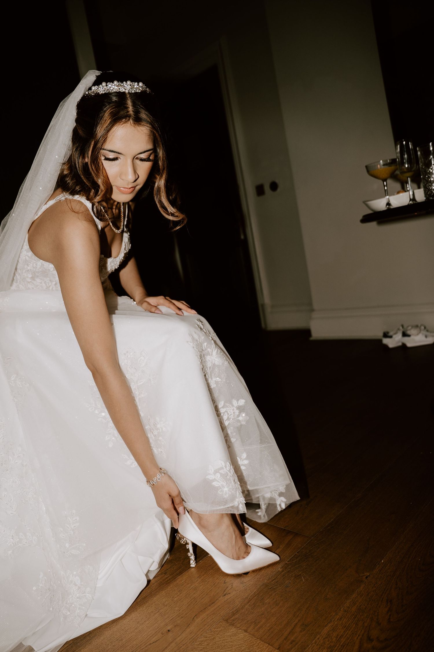 A bride in a white lace gown adjusts her shoes on a wooden floor.