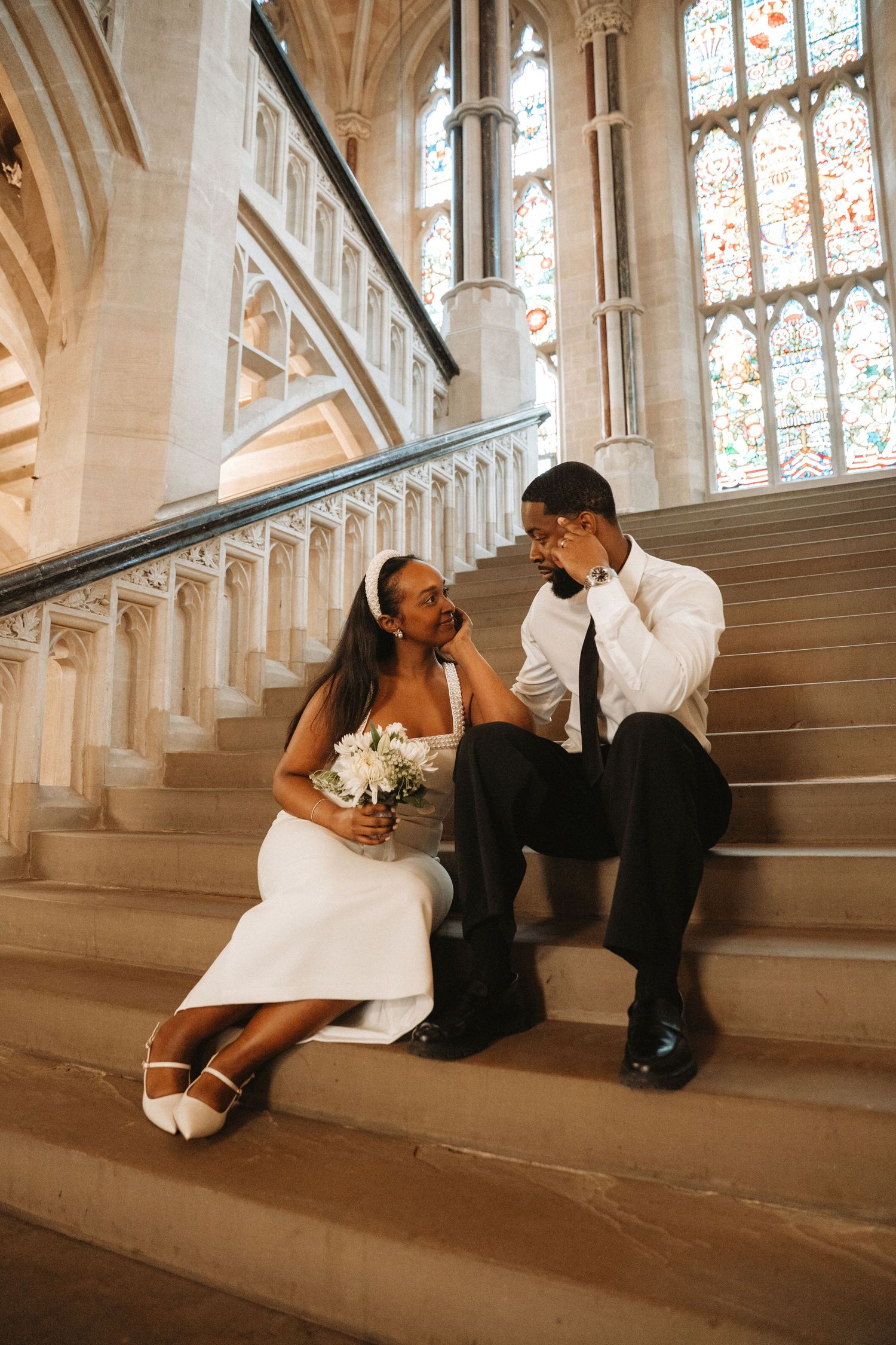 Bride and groom sit on church stairs, she holds white bouquet, stained glass windows behind them.