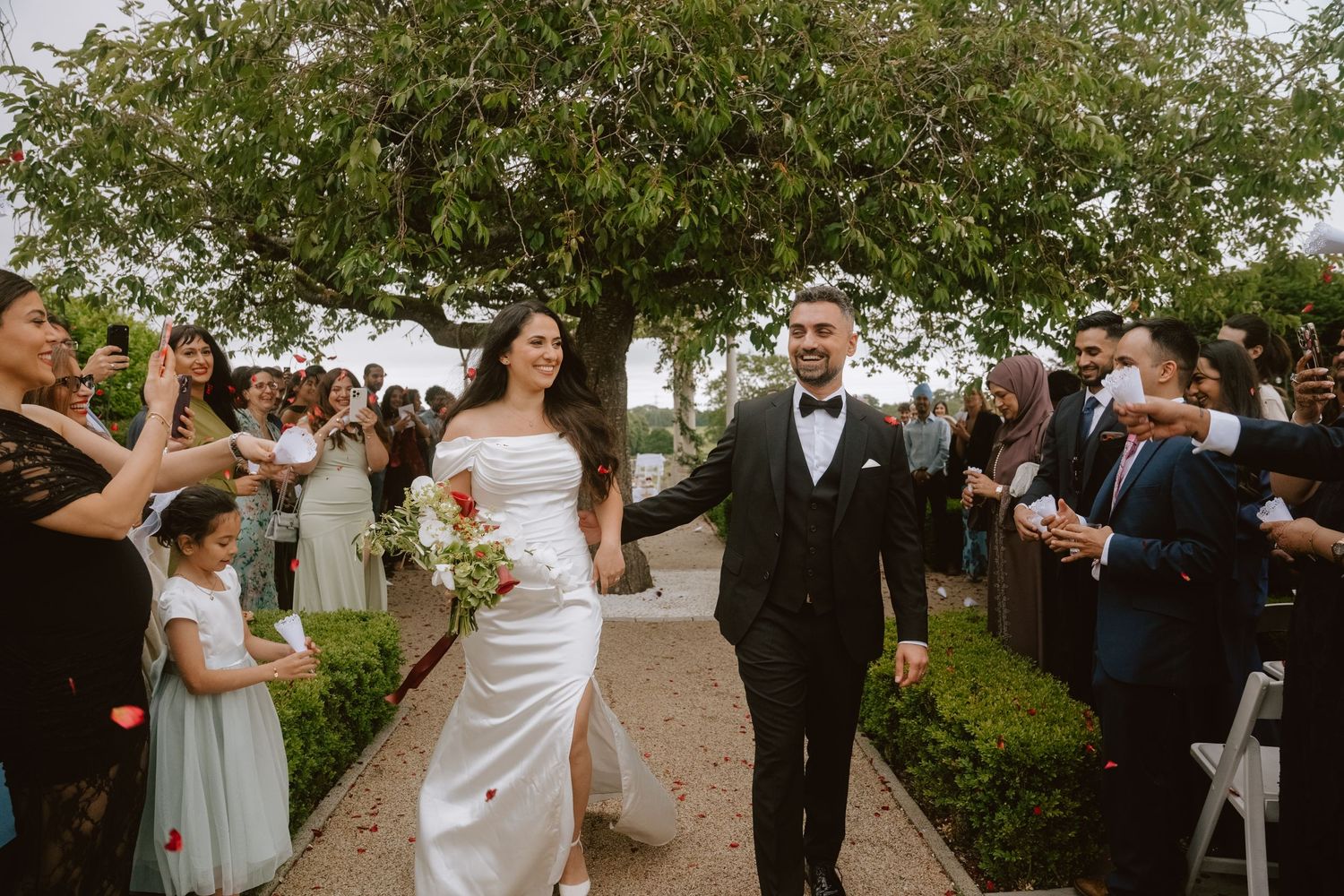 Bride and groom walk down outdoor wedding aisle while guests celebrate with confetti under lush green trees.
