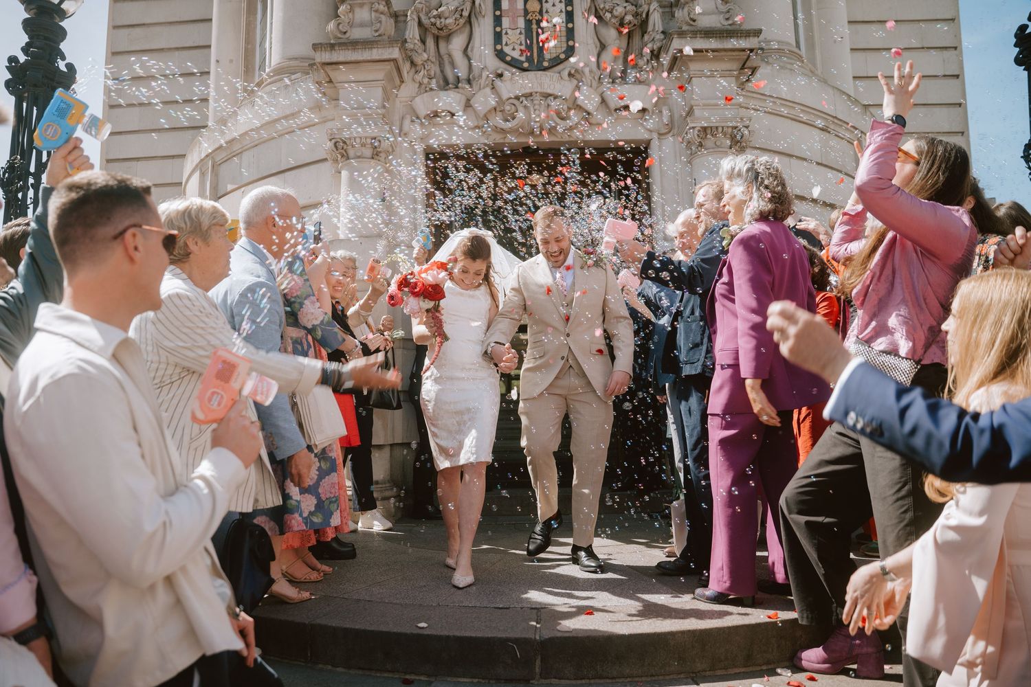 Newlywed couple exits building in confetti shower, surrounded by cheering guests at a sunny outdoor wedding celebration.