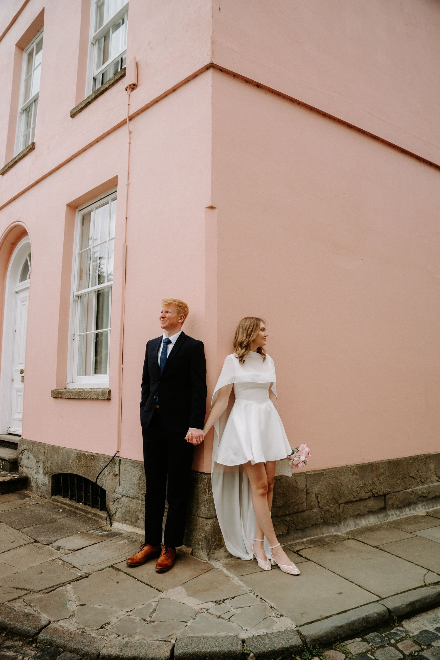 Bride and groom hold hands leaning against a pink building corner on cobblestone street, bride in short white dress.