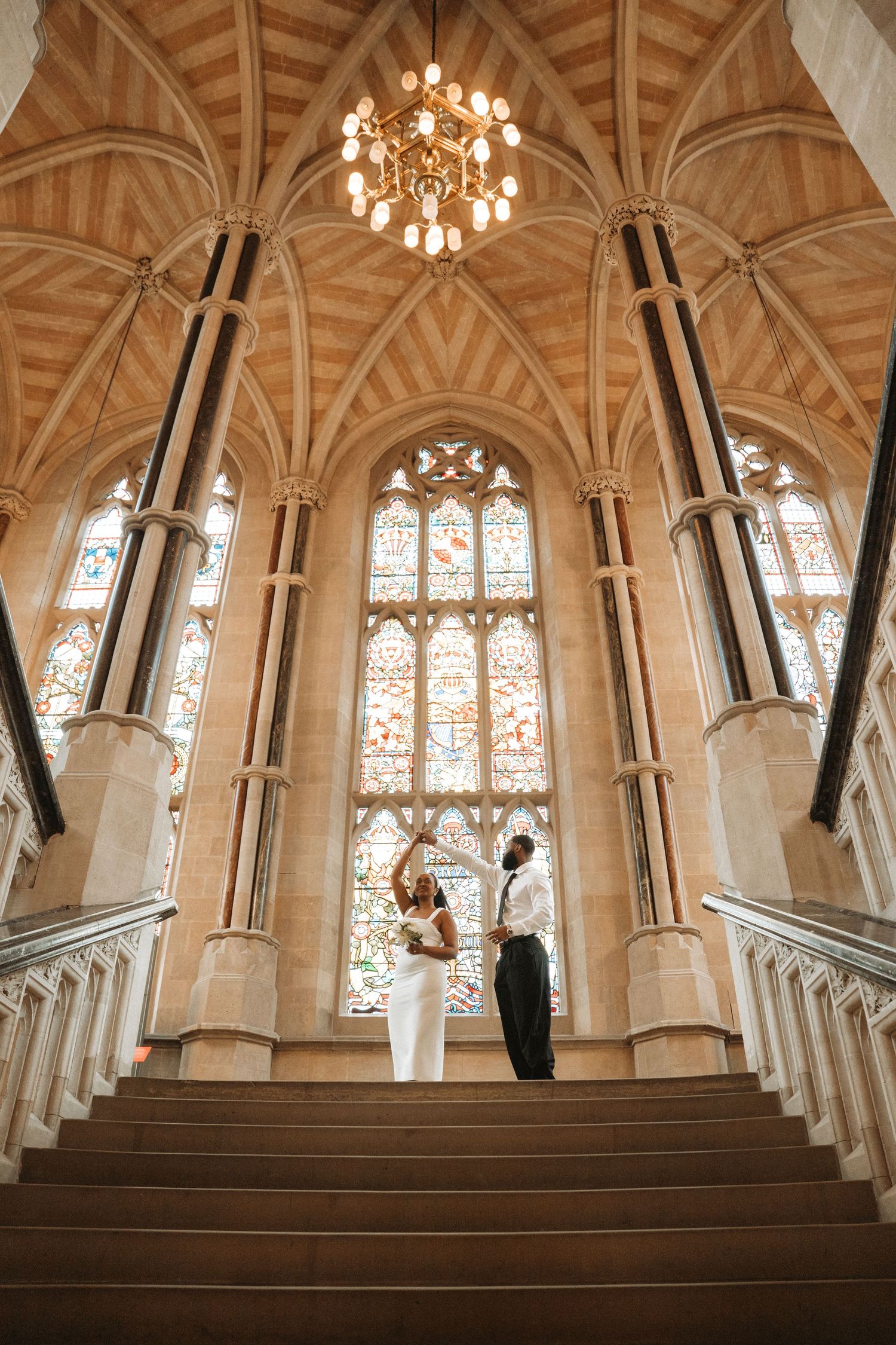 Bride and groom dancing on grand staircase beneath gothic arched ceiling and stained glass window in elegant church.