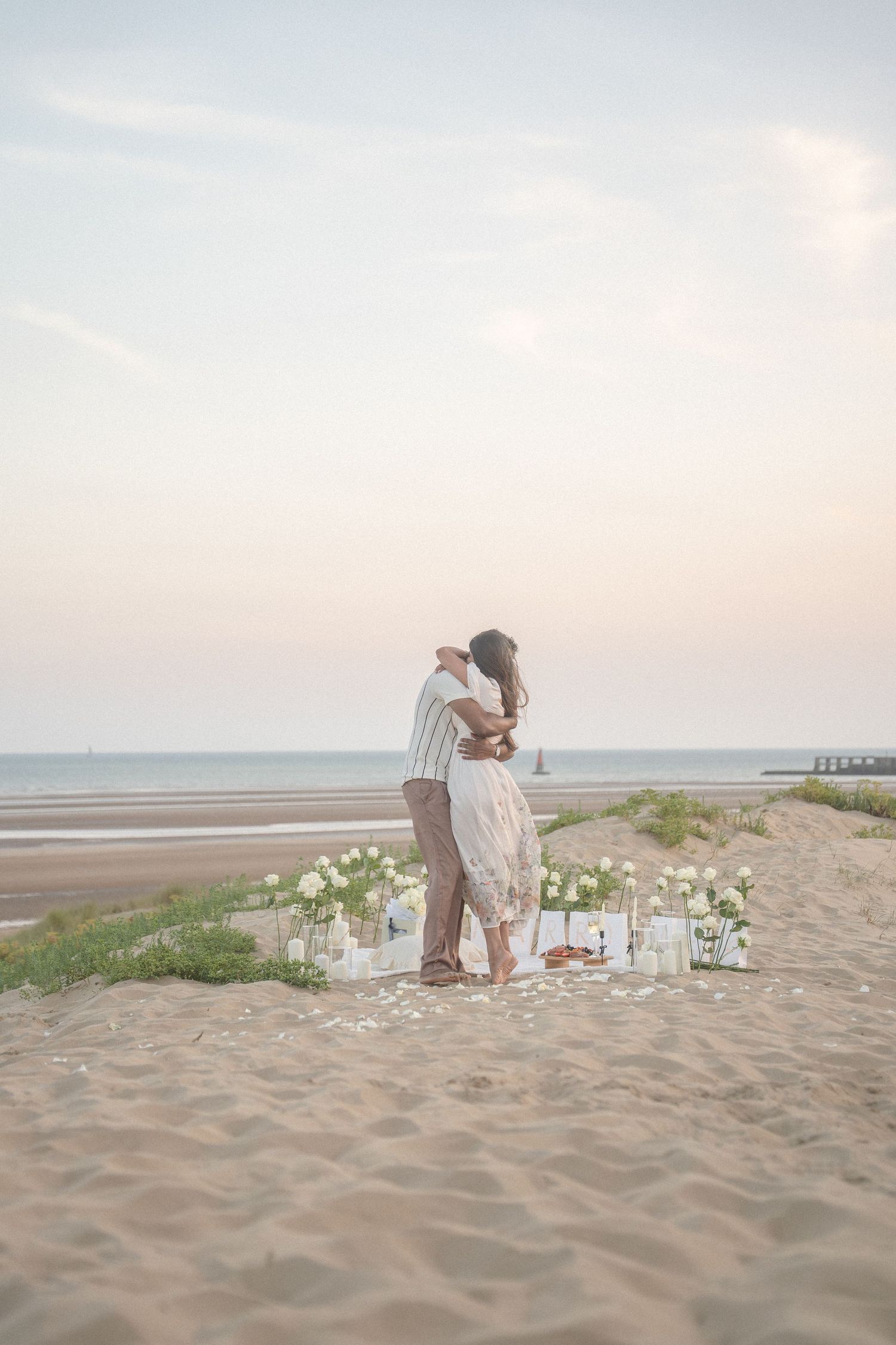 A romantic beach scene with candles and flowers as a couple embraces at sunset.