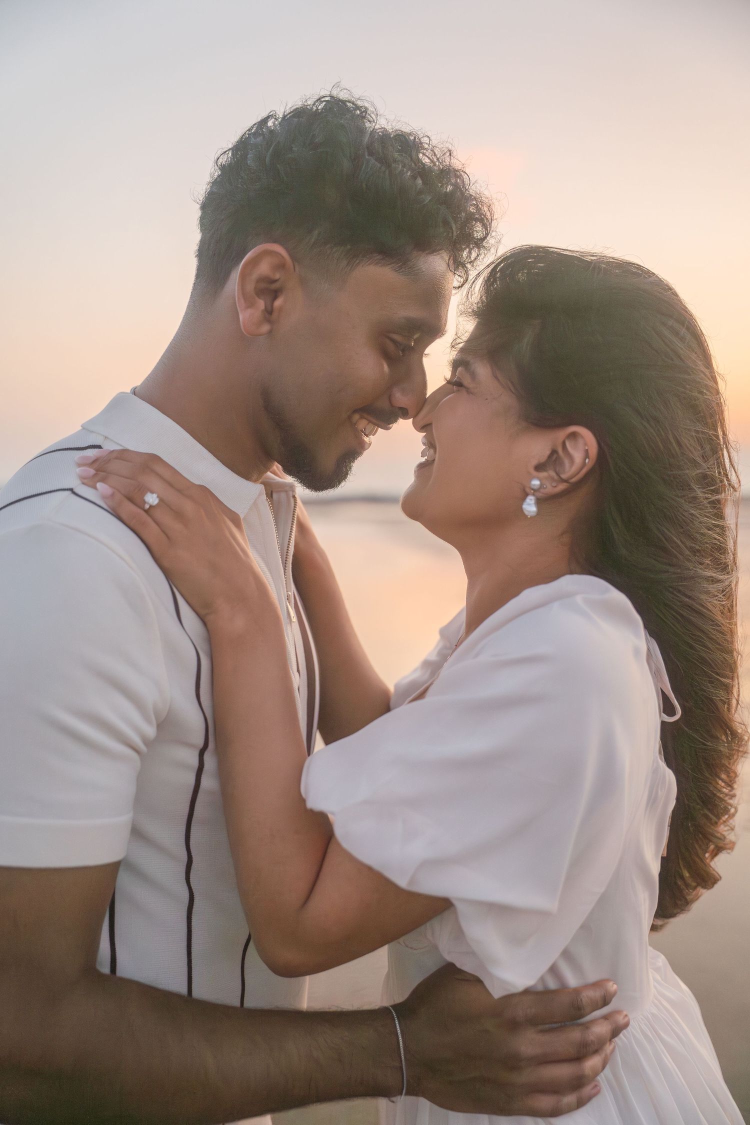 A tender romantic moment between a couple in white outfits against a sunset beach backdrop.