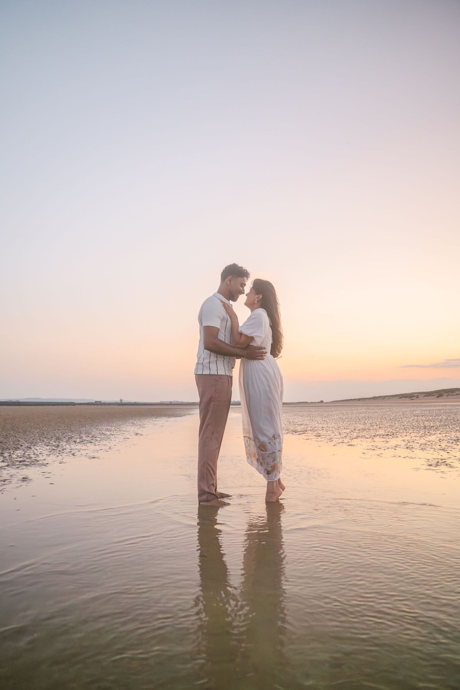 A romantic couple embraces while standing in shallow water during a beautiful sunset on the beach.