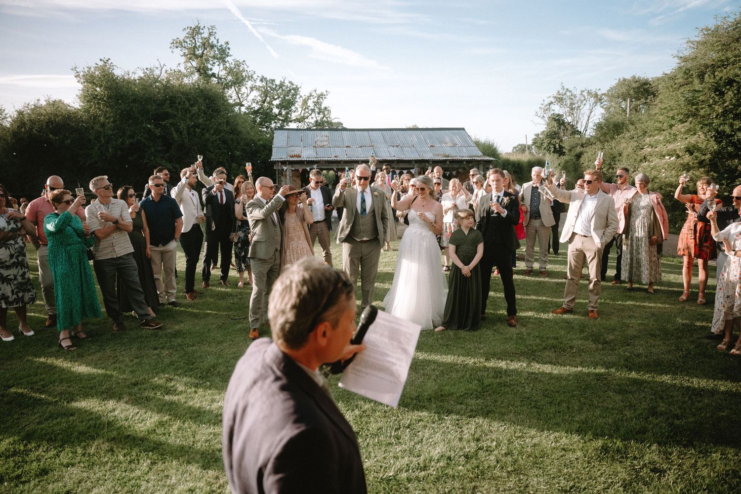 Outdoor wedding ceremony on green lawn with guests gathered around bride and groom while officiant reads from paper.
