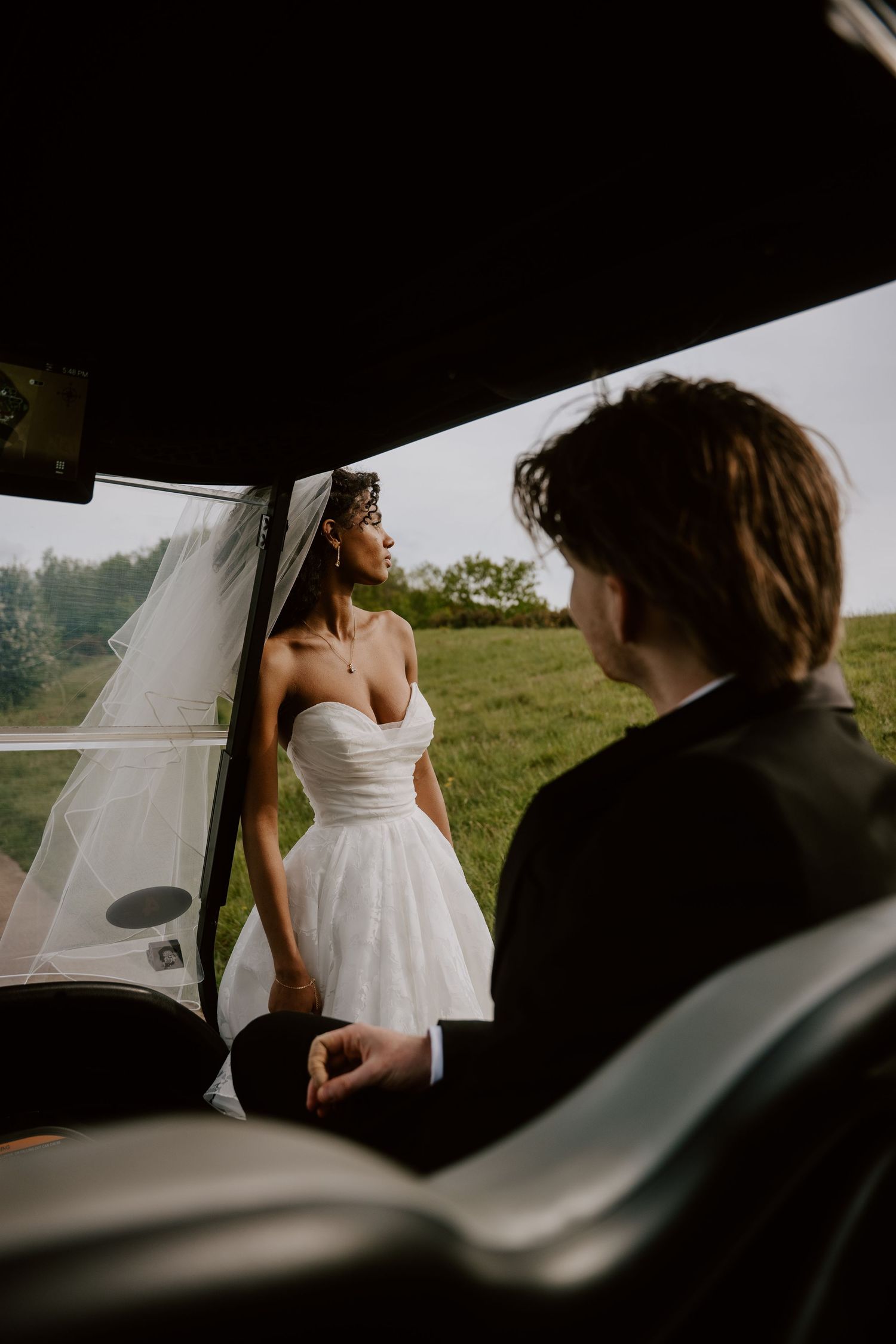 Bride in short white dress and veil stands outside vintage car as groom looks on, with green hills in background.