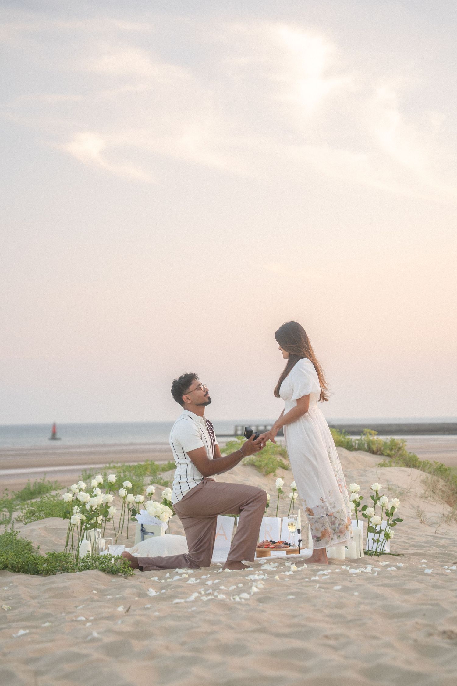 Beach proposal scene with white flowers and candles arranged on sandy dunes at sunset.
