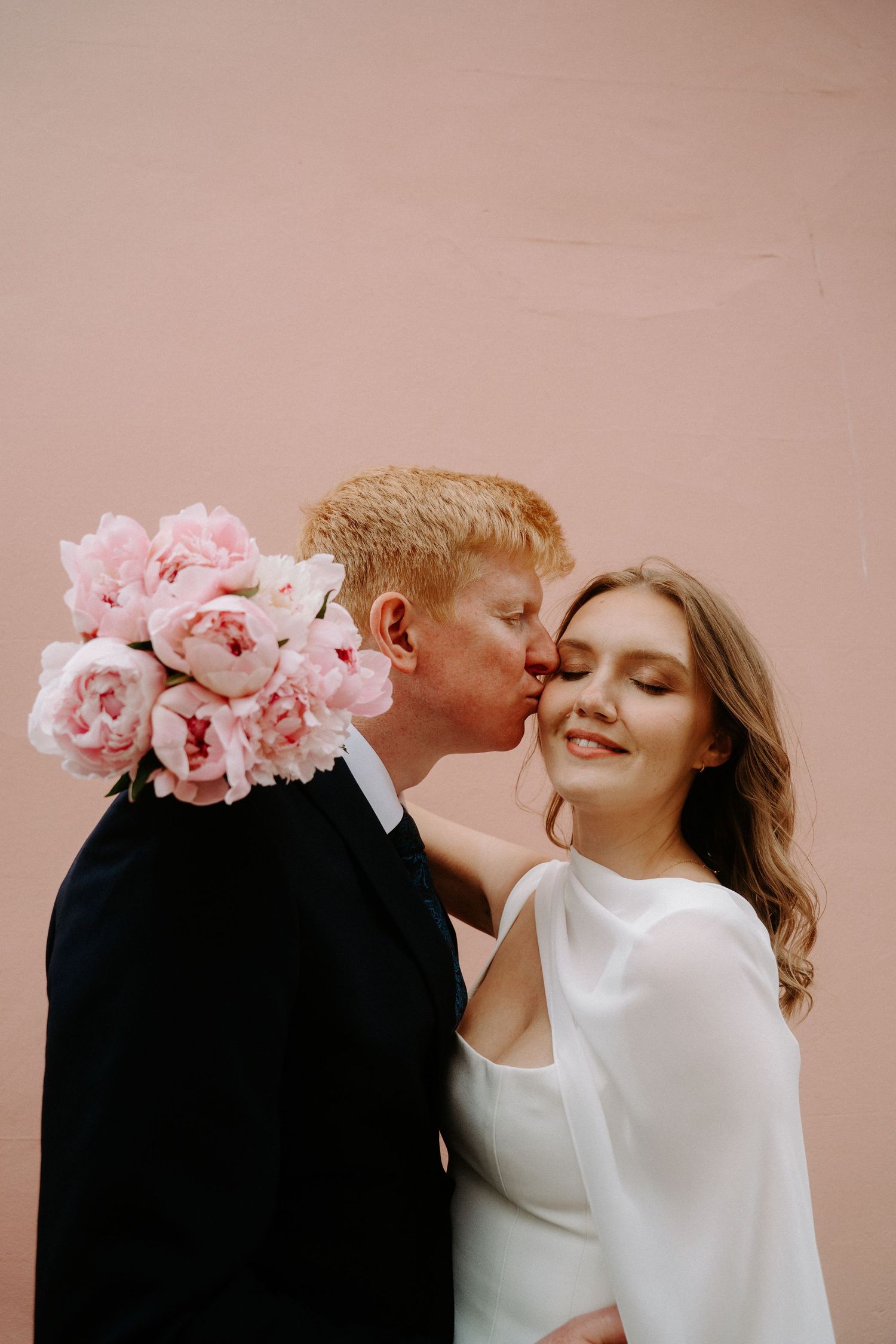 Groom in black suit kissing bride on cheek while holding pink peonies bouquet against a blush pink wall.
