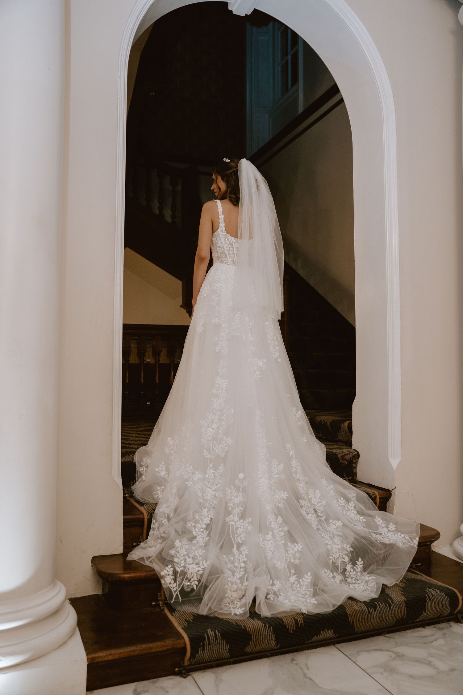 A flowing white wedding dress train cascades down a staircase with white architectural columns.
