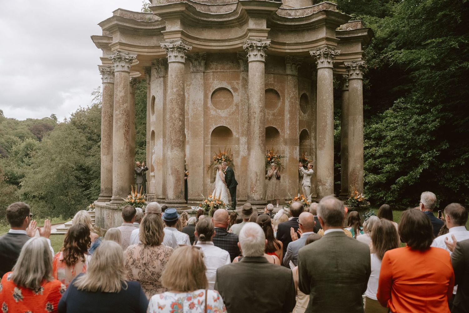 Outdoor wedding ceremony at ancient stone temple with columns, guests watching couple exchange vows surrounded by lush greenery.