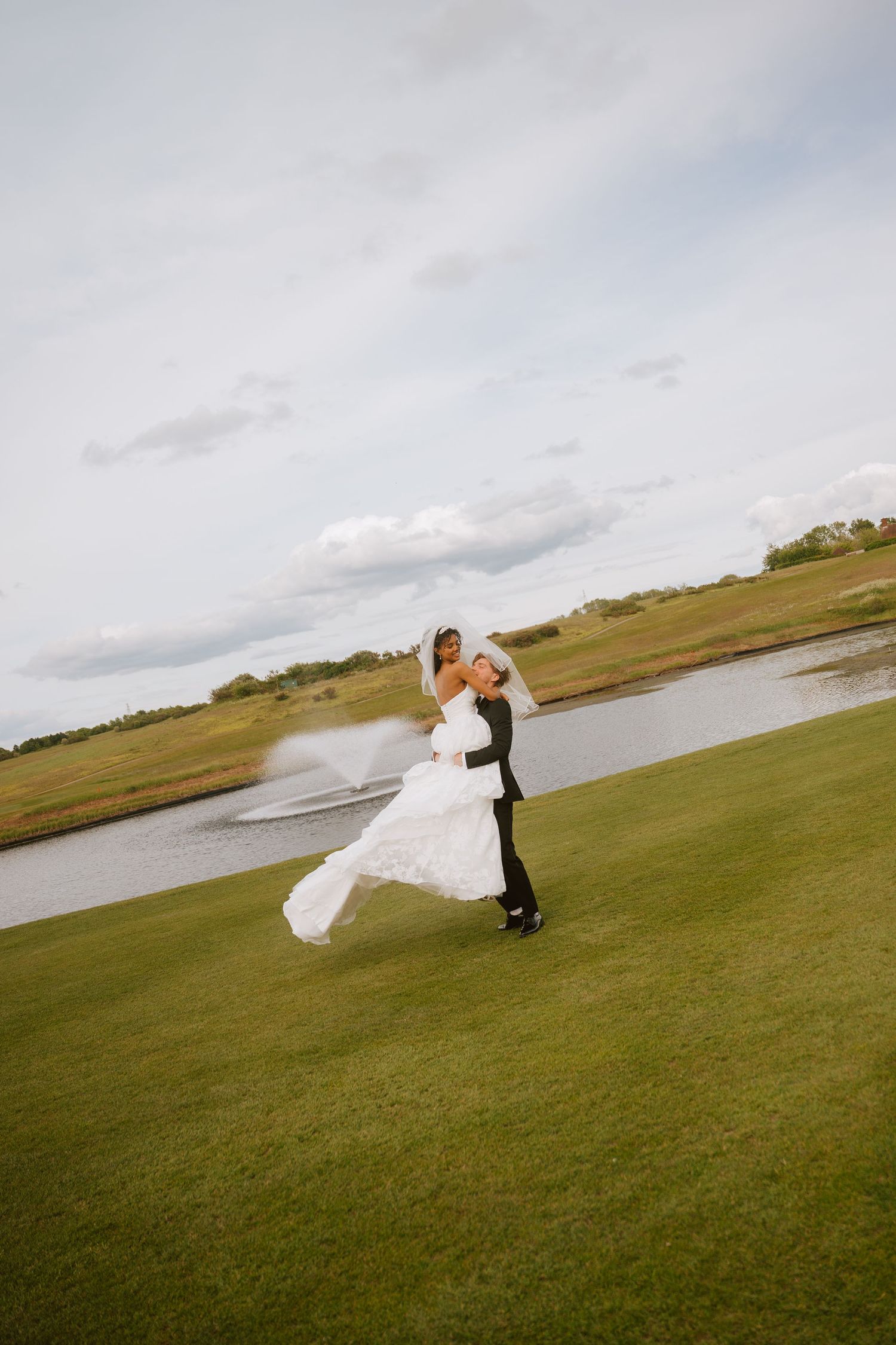 Groom lifting bride in white dress on lush green golf course with pond backdrop on their wedding day.