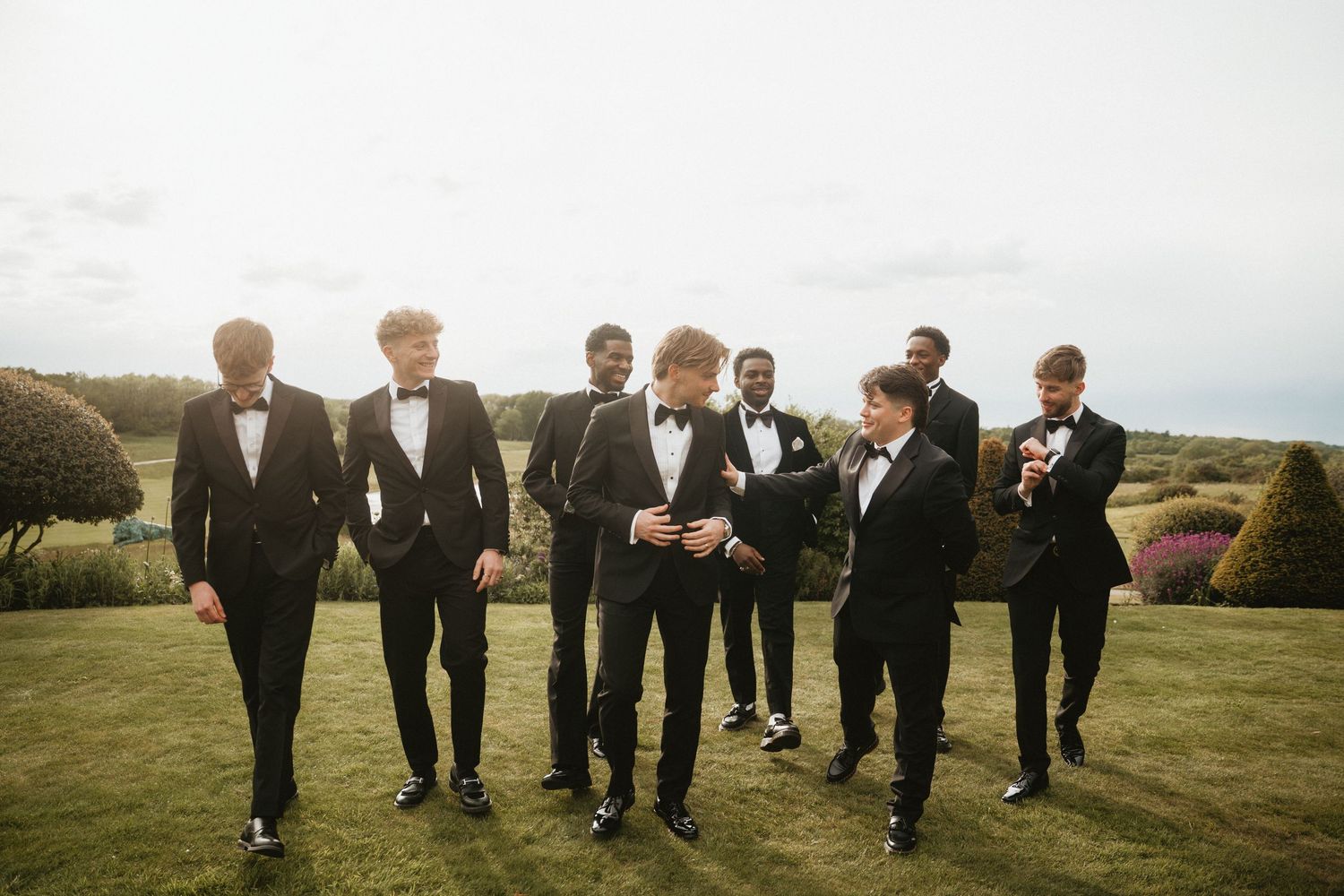 Seven men in black tuxedos and bow ties walk together on a green lawn at an outdoor wedding celebration.