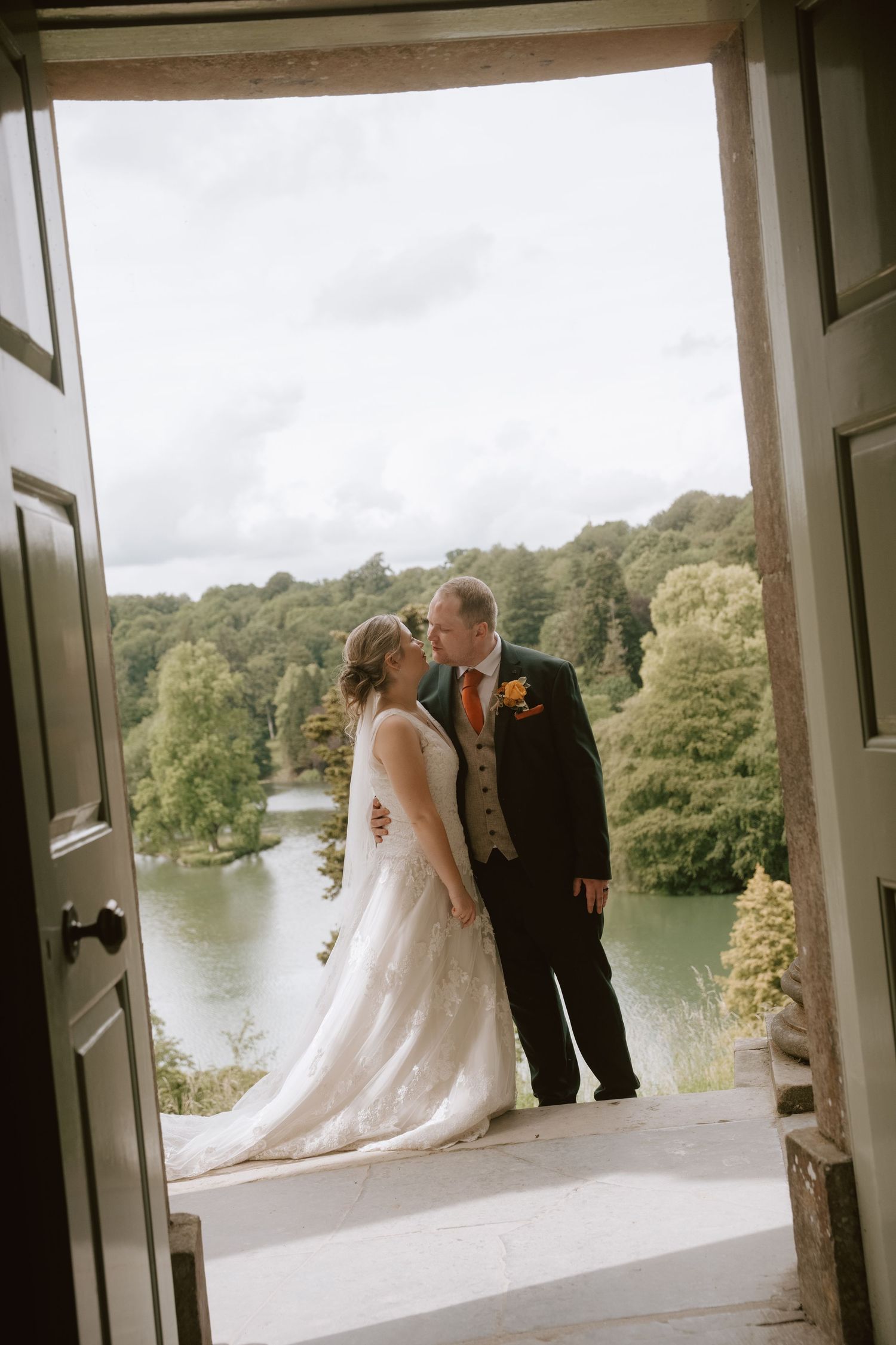 Bride and groom kissing in a doorway with a scenic lake and lush green trees in the background on their wedding day.
