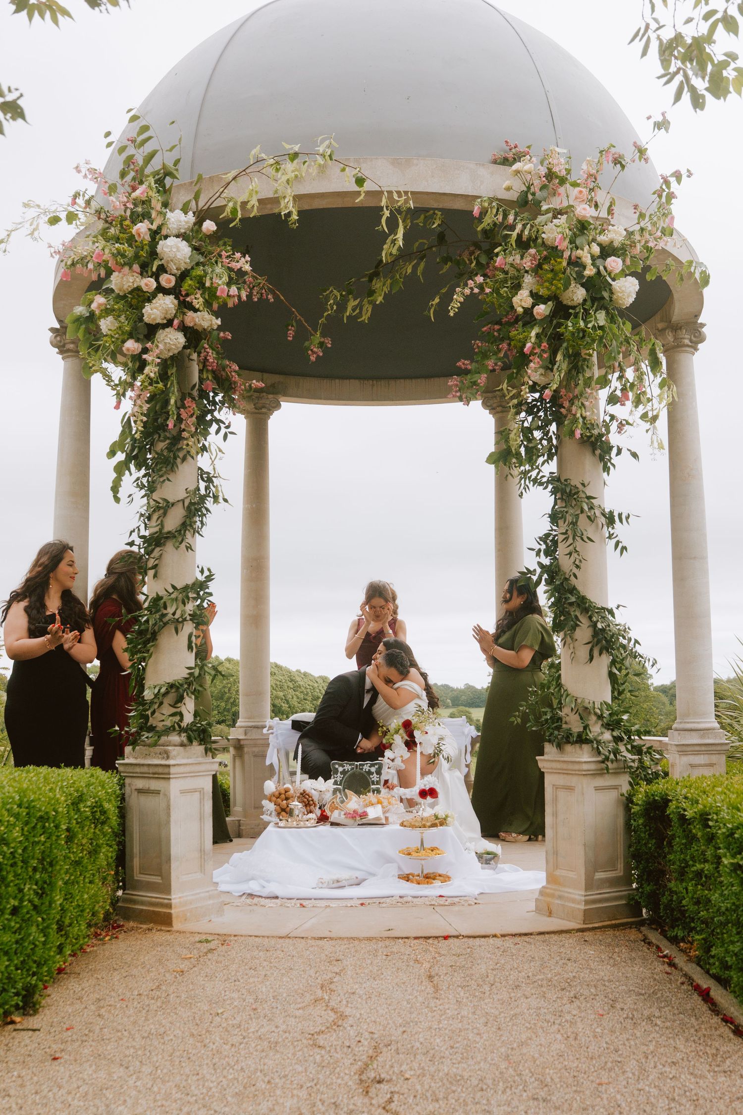 Couple cuts wedding cake under floral-adorned gazebo with bridesmaids watching at elegant outdoor garden ceremony.