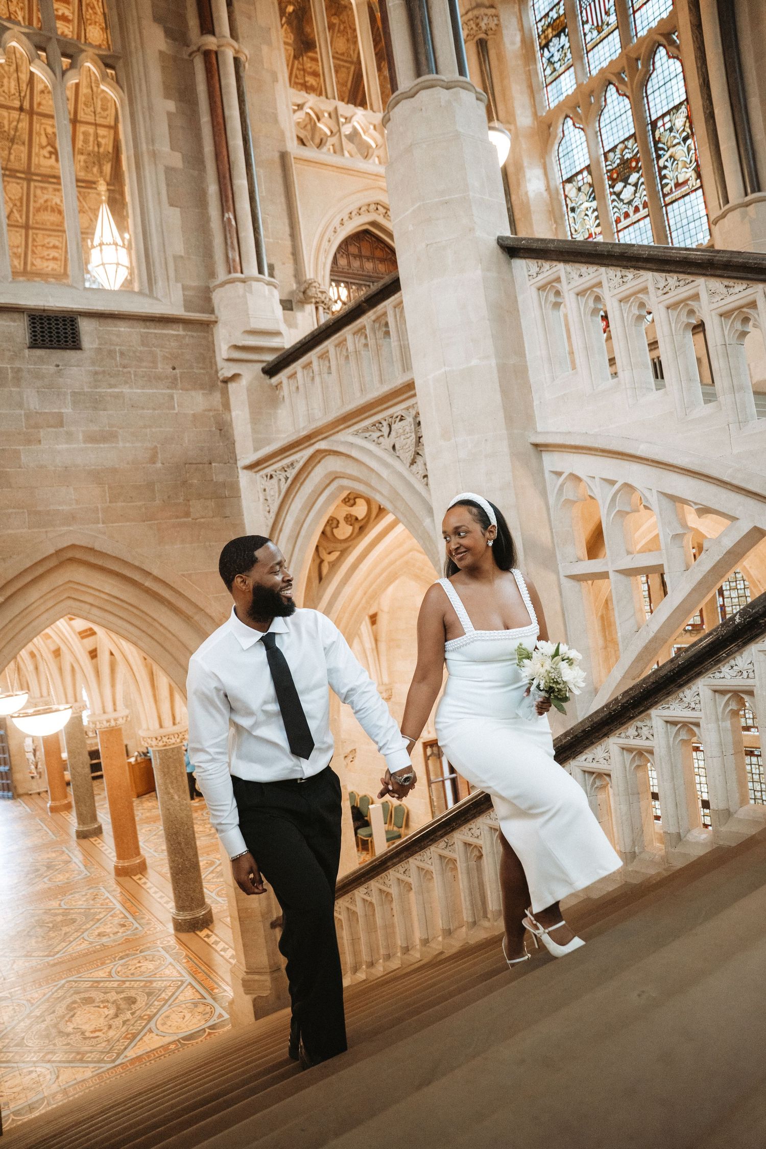 Bride and groom holding hands on ornate marble staircase in elegant gothic cathedral venue on wedding day.