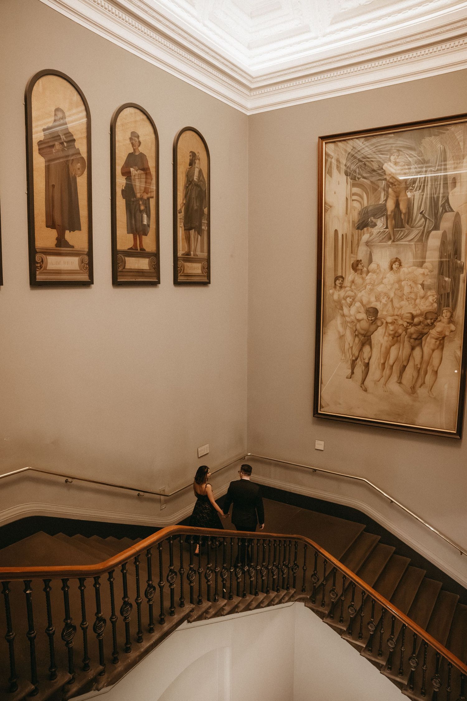 Vintage framed artwork and portraits line the walls of an elegant staircase with dark wooden railings and cream walls.